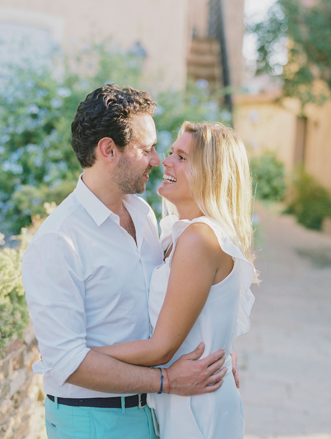 Un couple s'enlace et se sourit en plein air, dans un village méditerranéen, vêtu de blanc et de turquoise.