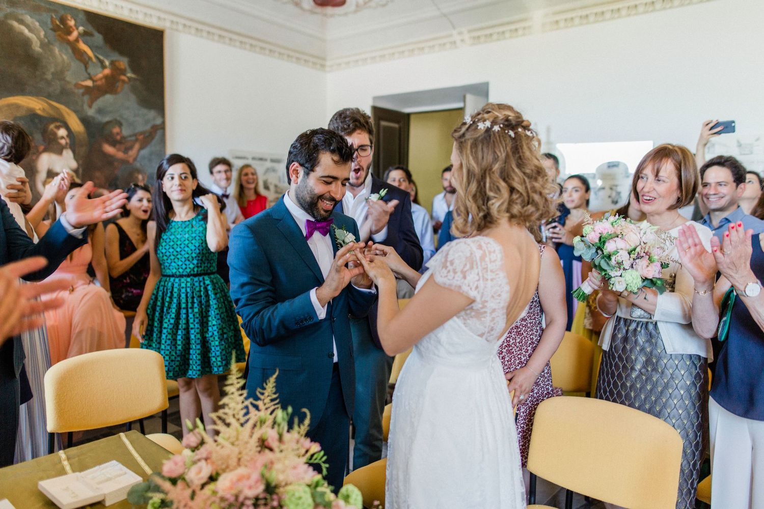 Bride and groom exchange rings during wedding ceremony while guests applaud in elegant venue.