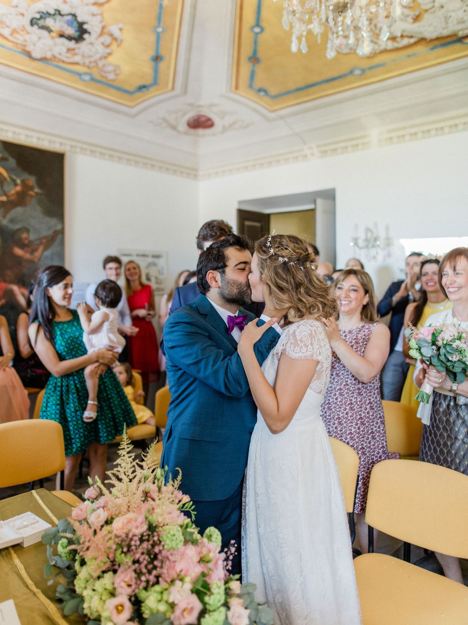 Bride and groom share first kiss at elegant wedding ceremony in ornate room with guests celebrating.