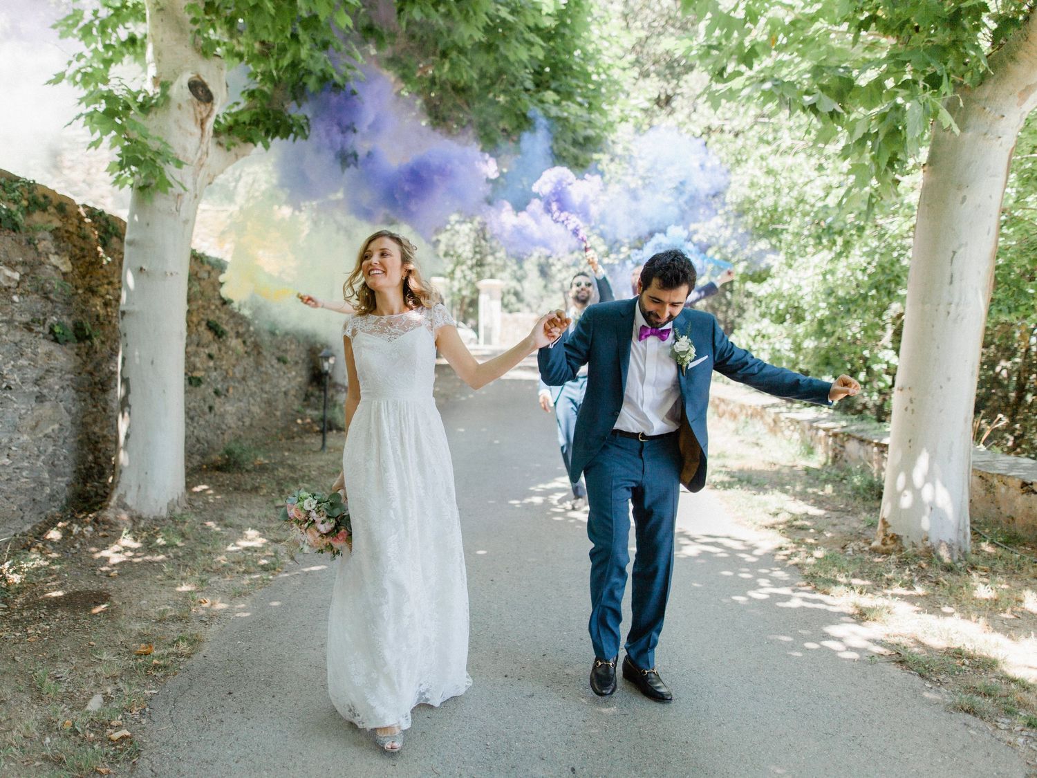 Joyful newlyweds walk hand-in-hand holding colorful purple smoke bombs on a tree-lined path in a wedding celebration.