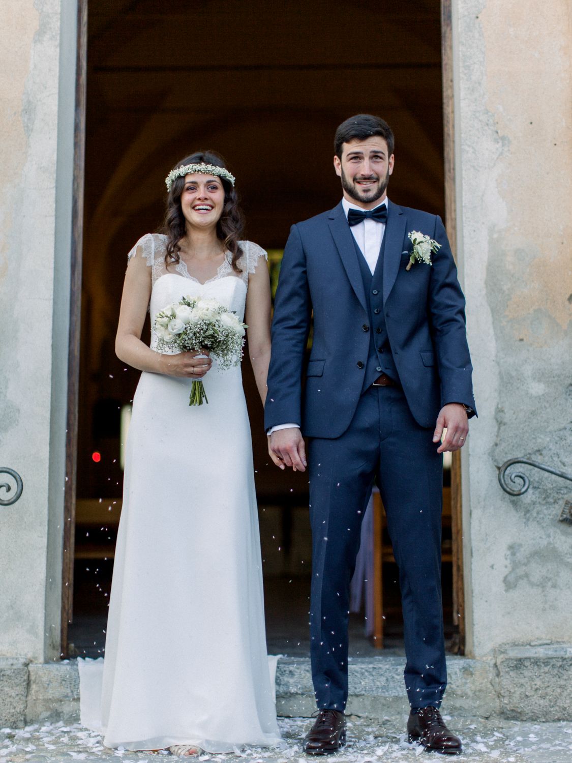Smiling bride and groom holding hands outside church, bride in white gown with floral crown, groom in navy suit.