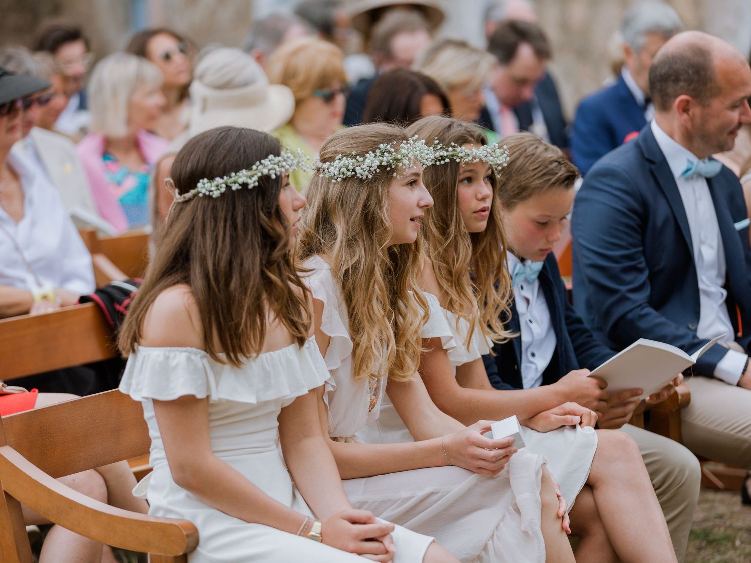 De jeunes filles en robes blanches et couronnes de fleurs sont assises sur des bancs en bois lors d'une cérémonie de mariage en plein air.