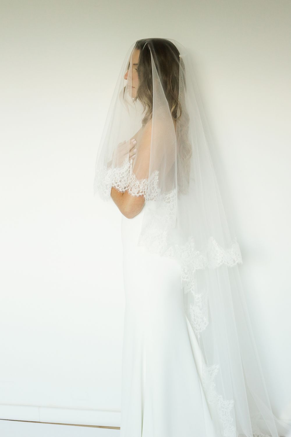 Bride in a white lace wedding gown and veil draped over her head, standing elegantly against a light wall.