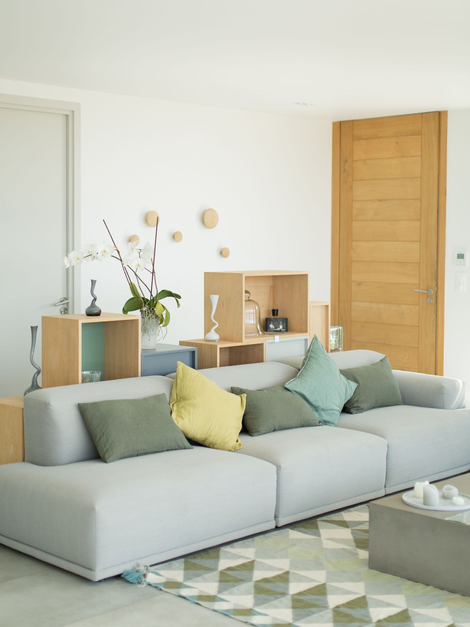 Salon moderne avec canapé modulable gris clair, coussins colorés, étagère en bois et tapis à motifs géométriques.
Maisons de Julia.