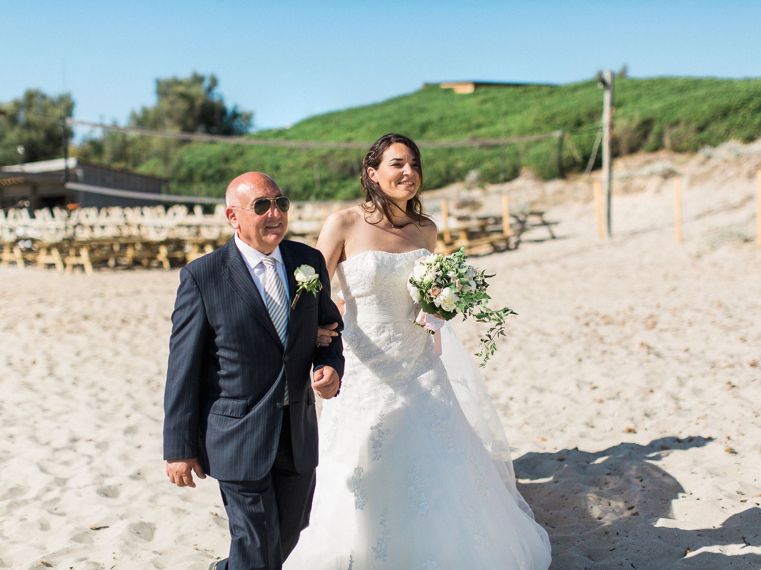 La mariée en robe blanche marche avec un homme plus âgé sur la plage de sable lors d'une cérémonie de mariage en plein air.