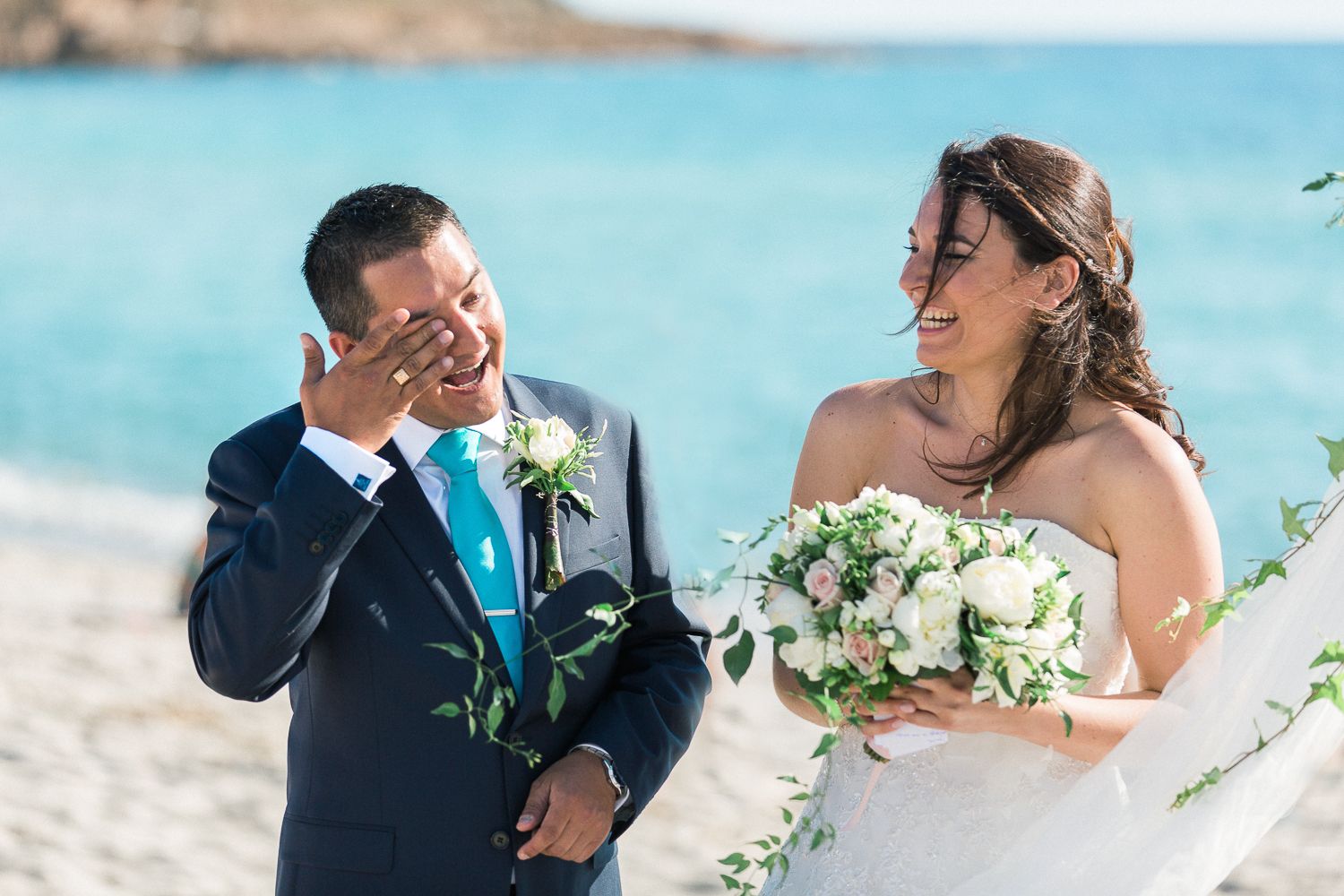 Le marié, ému, essuie ses larmes à côté de la mariée souriante qui tient un bouquet blanc lors d'une cérémonie de mariage sur la plage.