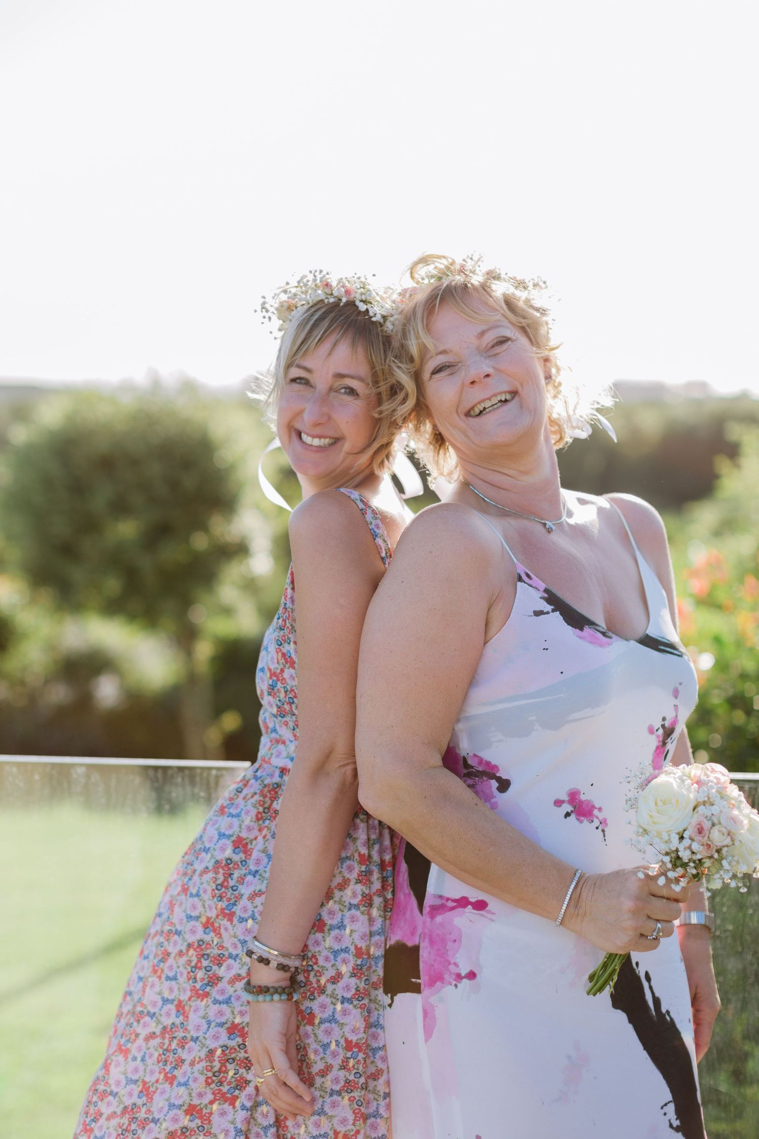 Deux femmes souriantes en robes d'été posent dos à dos à l'extérieur, l'une tenant un petit bouquet blanc.