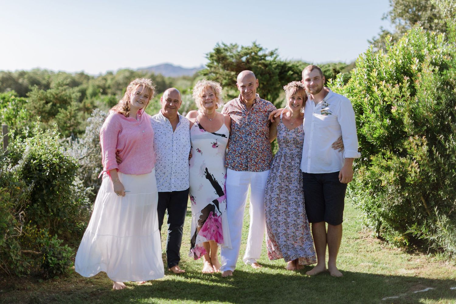 Un groupe de six adultes, visiblement heureux, pose ensemble en extérieur dans un jardin verdoyant et luxuriant par une journée ensoleillée.