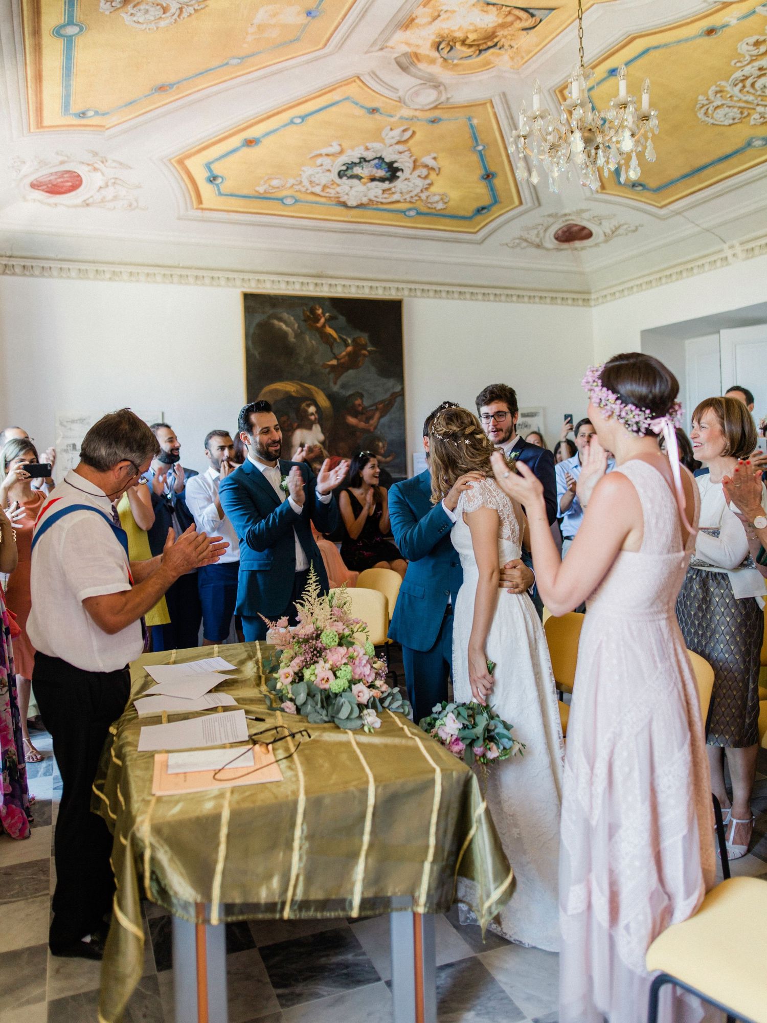 Elegant wedding ceremony in ornate room with frescoed ceiling, bride holding bouquet, guests applauding.
