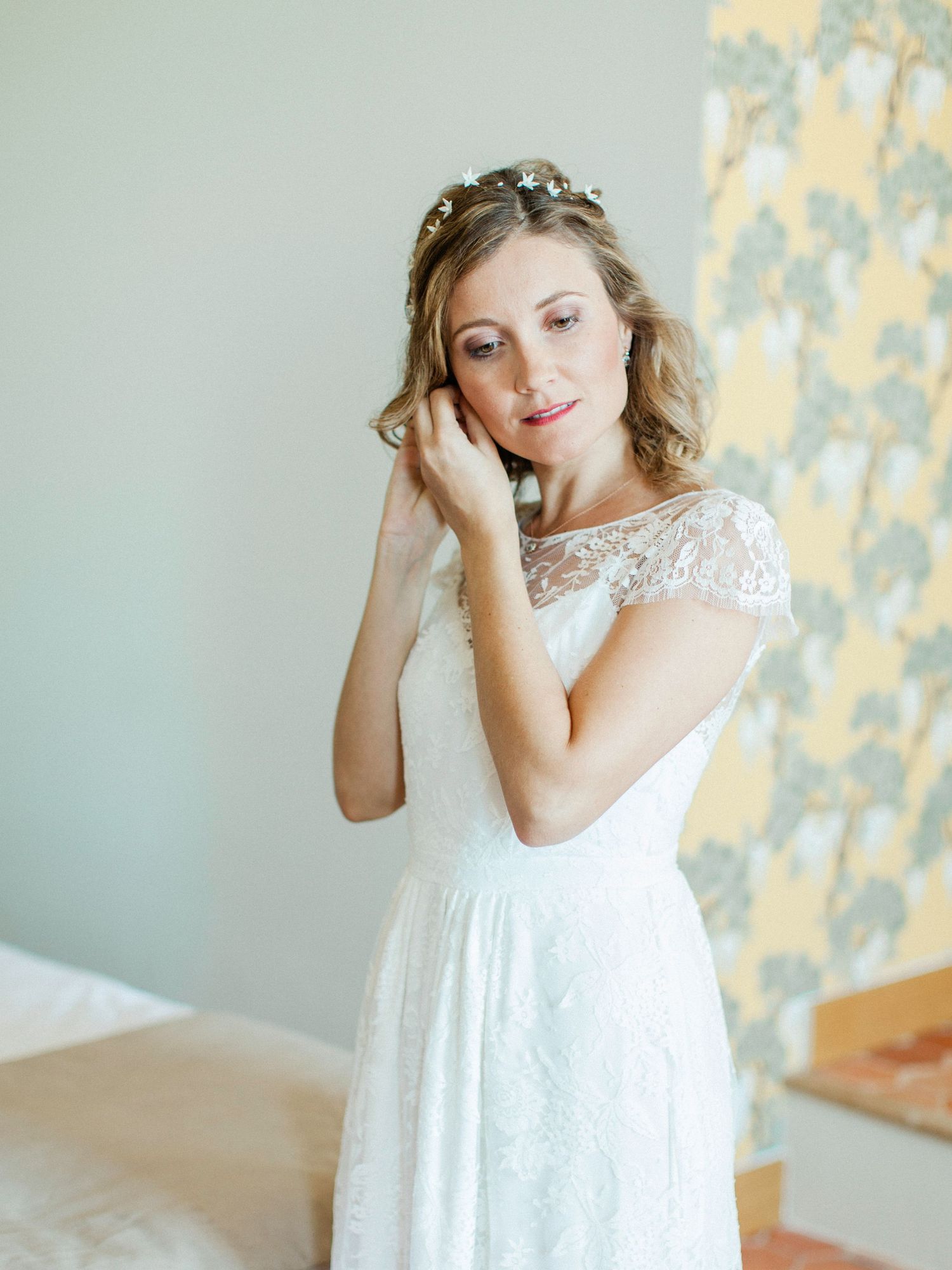 Bride in lace wedding dress adjusting earring, wearing floral headpiece, soft natural light indoor setting.