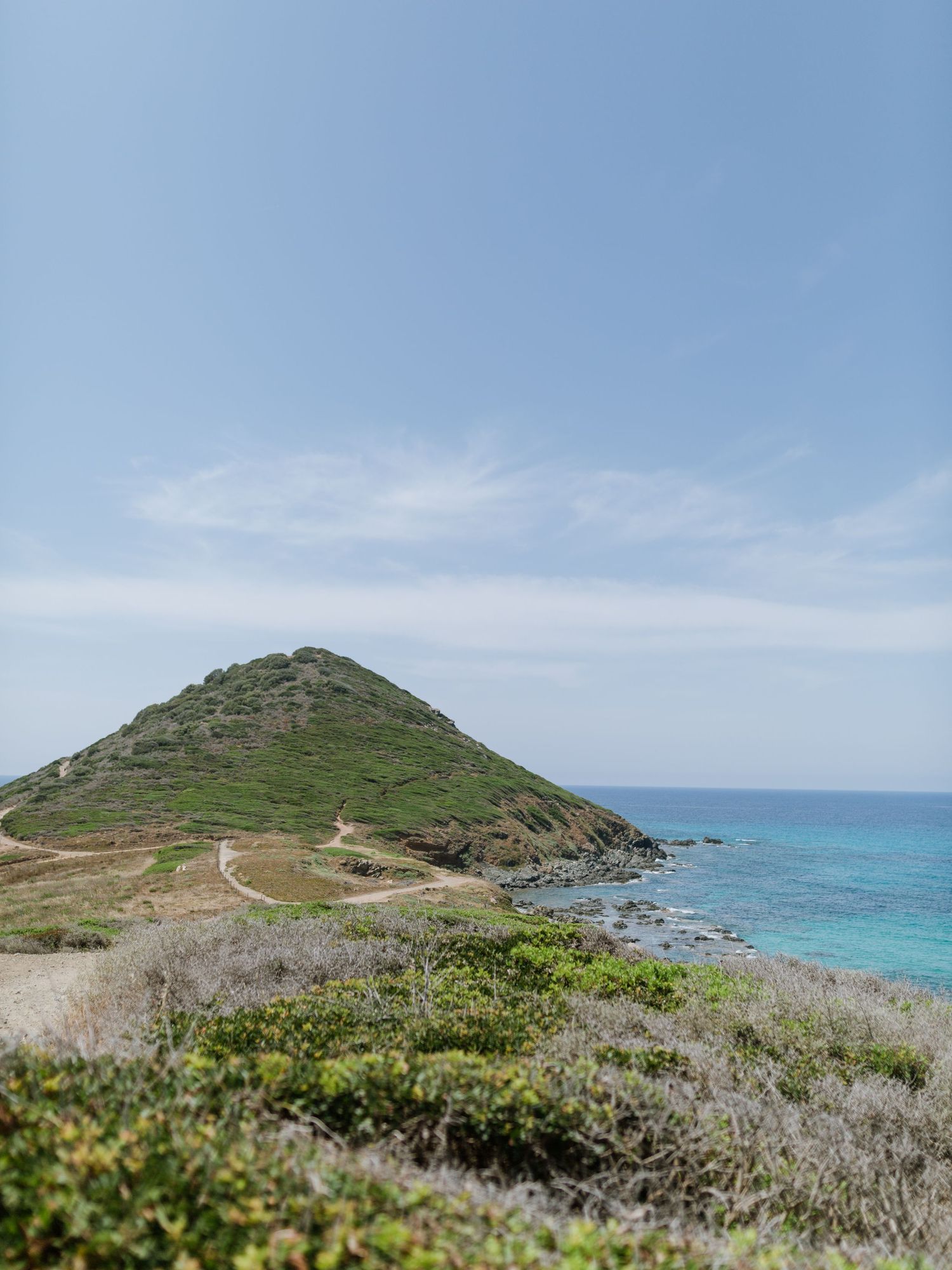 Un paysage côtier pittoresque avec une colline verdoyante, un sentier sinueux et un océan turquoise sous un ciel bleu limpide.
