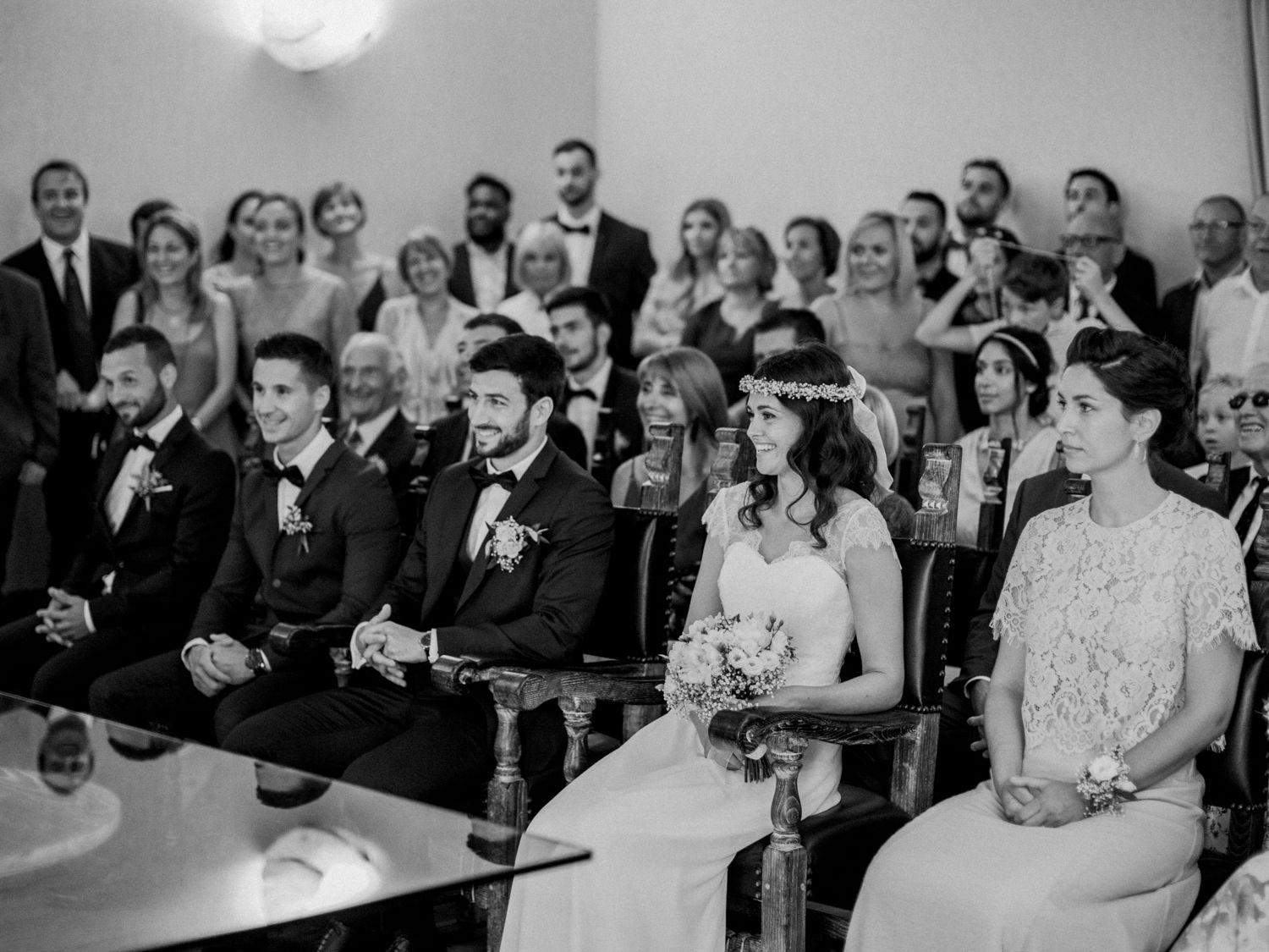 Black and white wedding ceremony photo with bride and groom seated alongside guests in elegant formal attire.