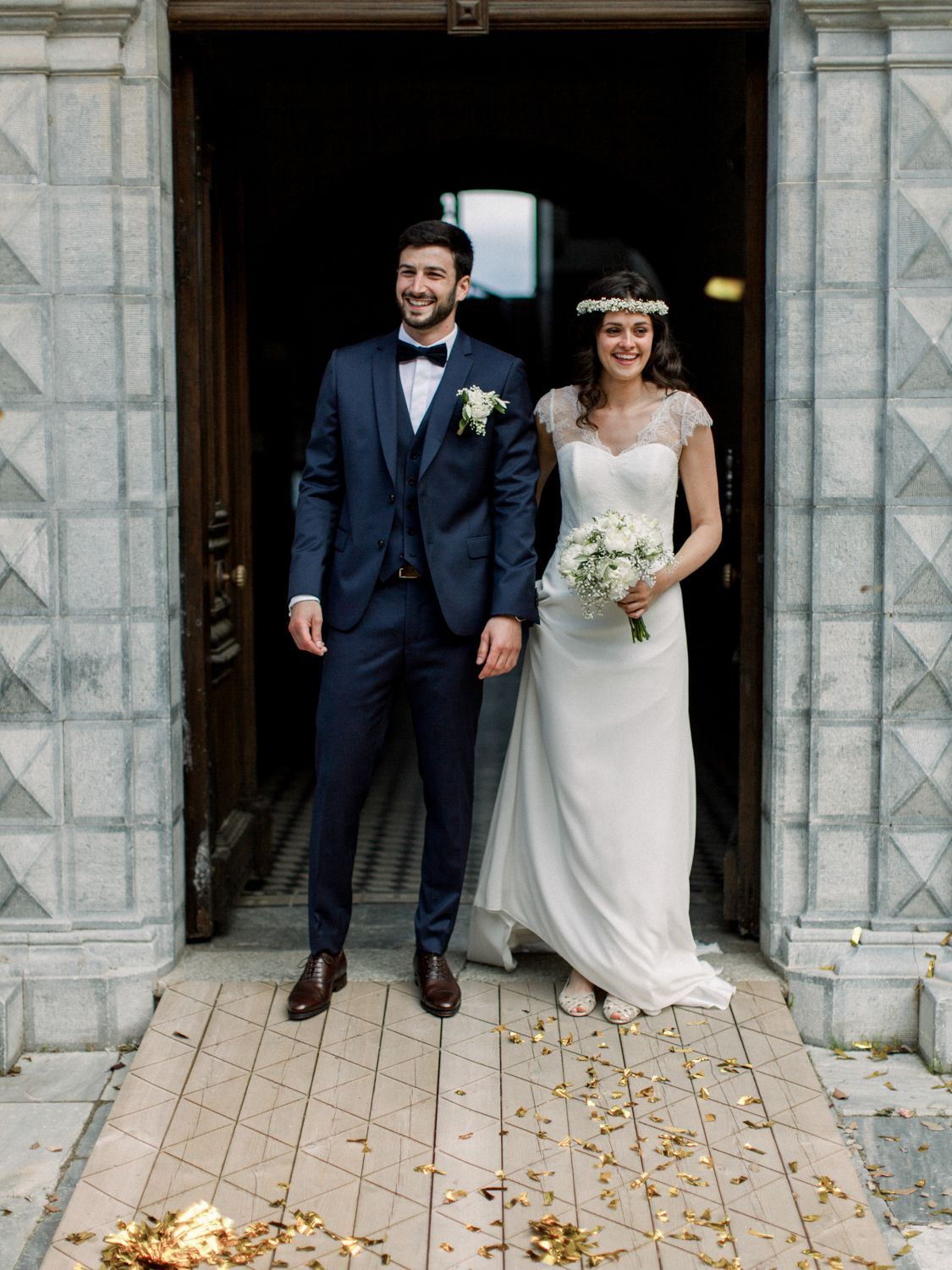 Smiling bride and groom stand at church entrance, bride holds white bouquet, groom wears navy suit with boutonniere.