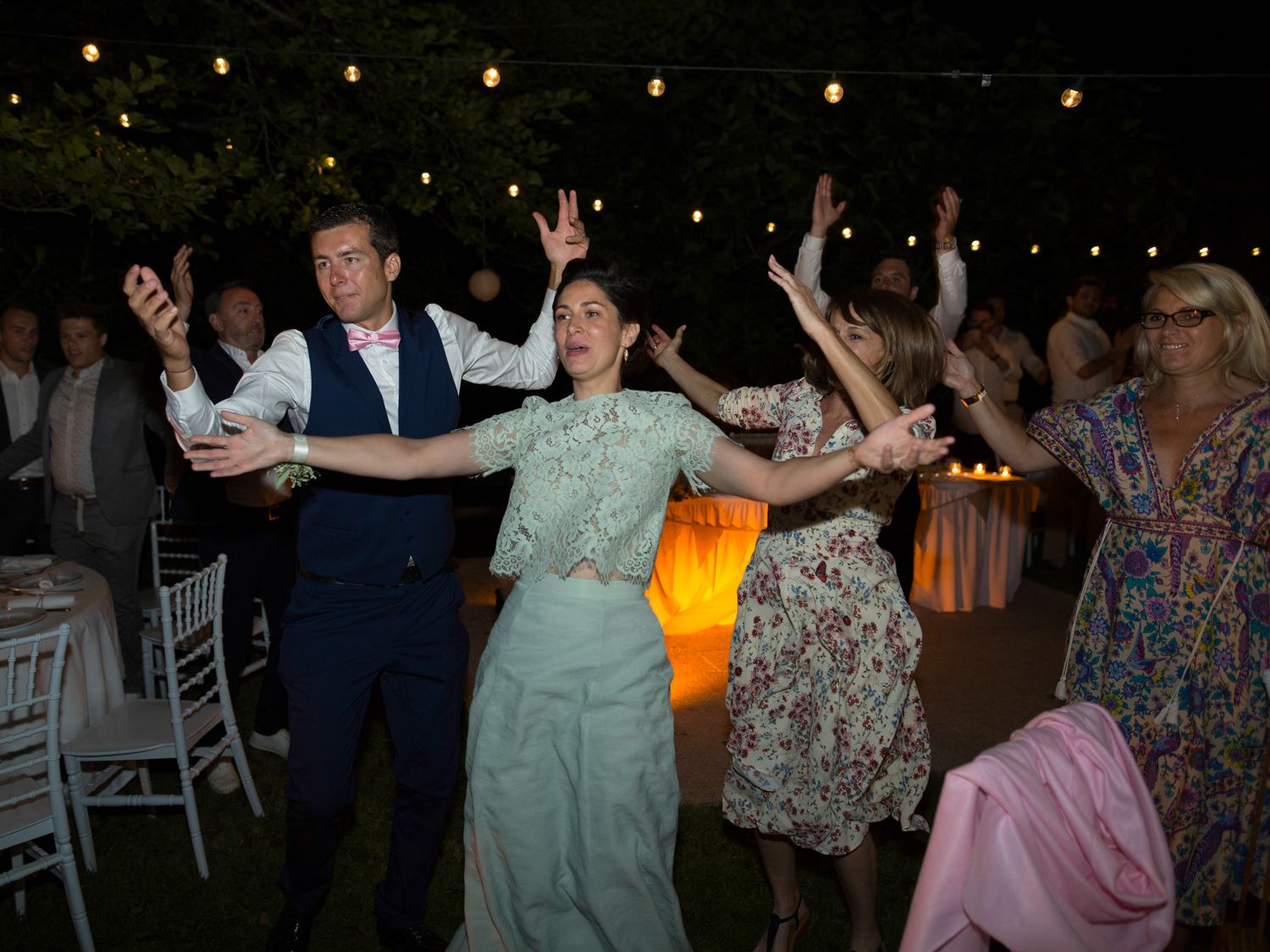 Des invités dansent joyeusement lors d'une réception de mariage en plein air, sous des guirlandes lumineuses.