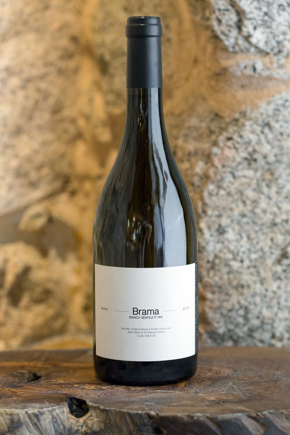 Bouteille de vin blanc Brama présentée sur une surface en bois rustique, devant un mur en pierre naturelle.