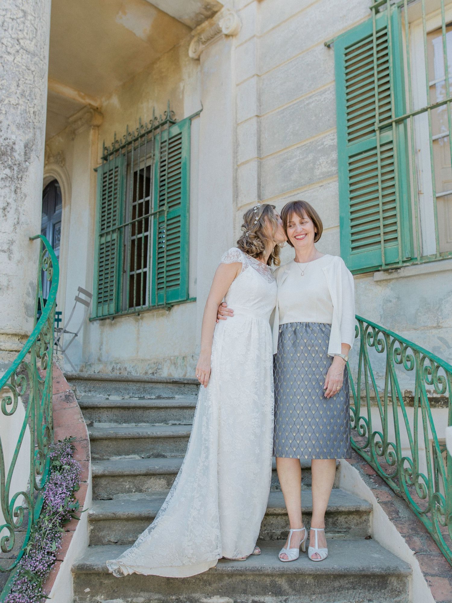 Bride in lace gown shares tender moment with guest on ornate steps of vintage villa with green shutters.