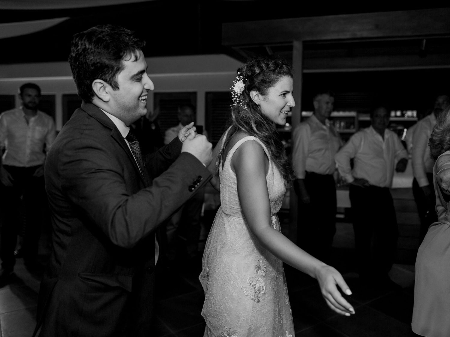 Photo en noir et blanc d'une mariée et d'un marié dansant joyeusement lors de leur réception de mariage, avec des invités en arrière-plan.