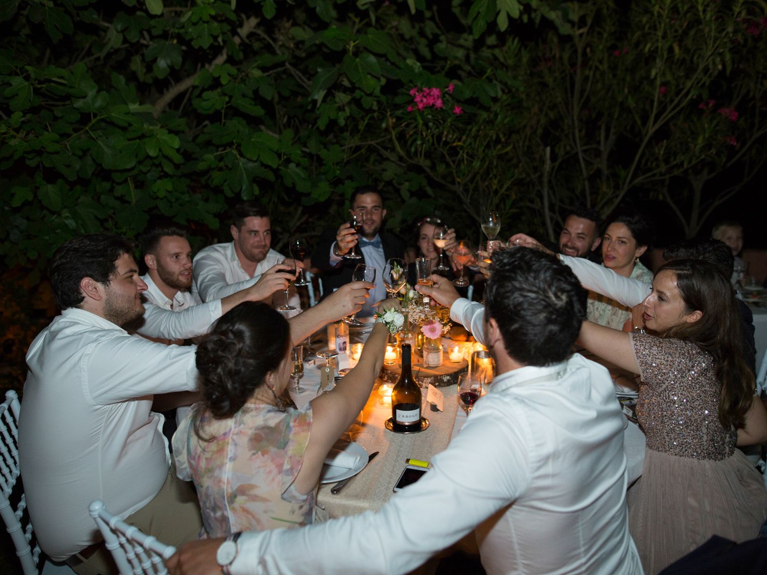 Un groupe d'invités élégamment vêtus trinquent lors d'un dîner en plein air sous une végétation luxuriante.