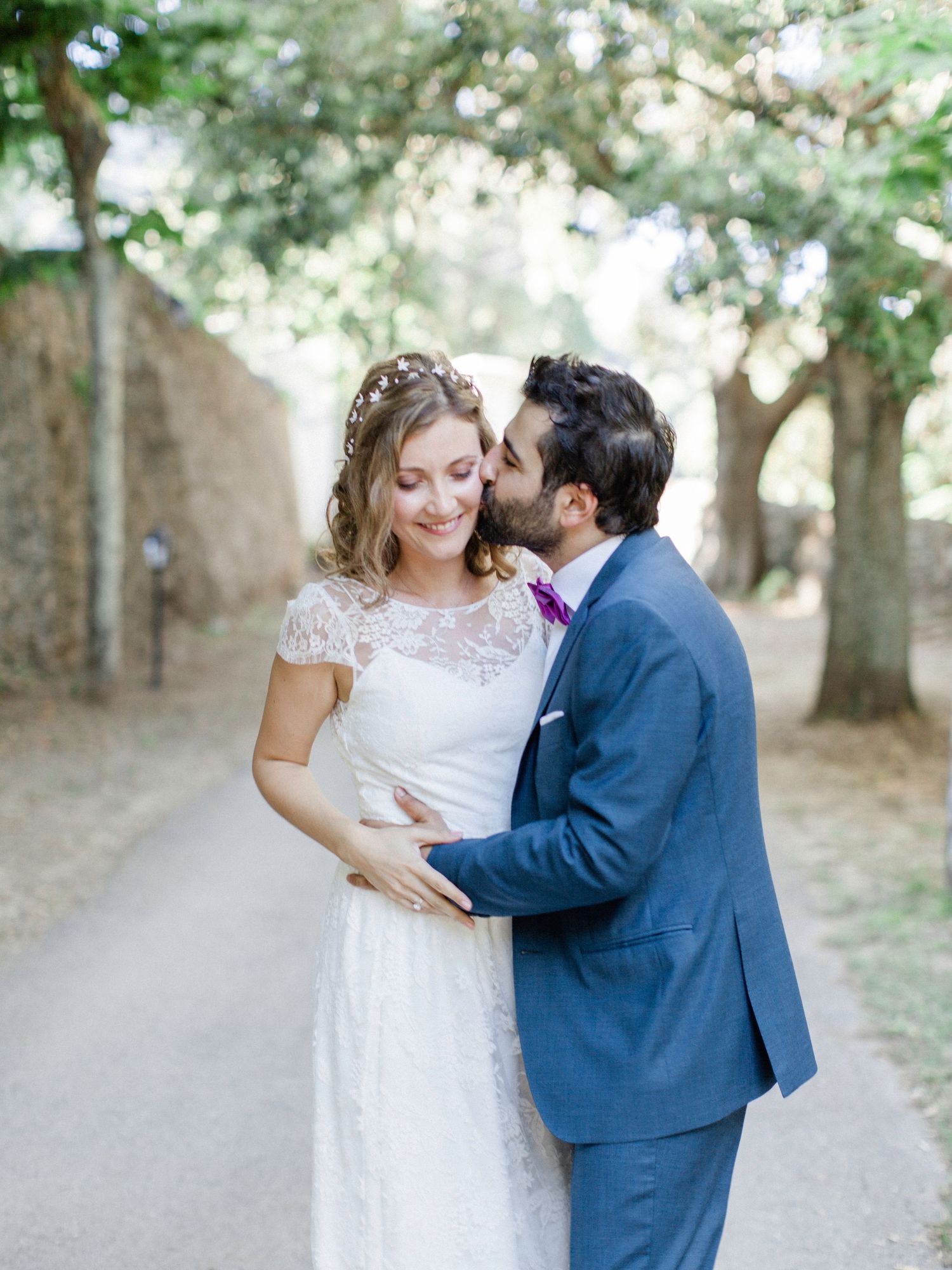 A groom in a blue suit kisses his bride in a lace wedding dress outdoors under green trees.