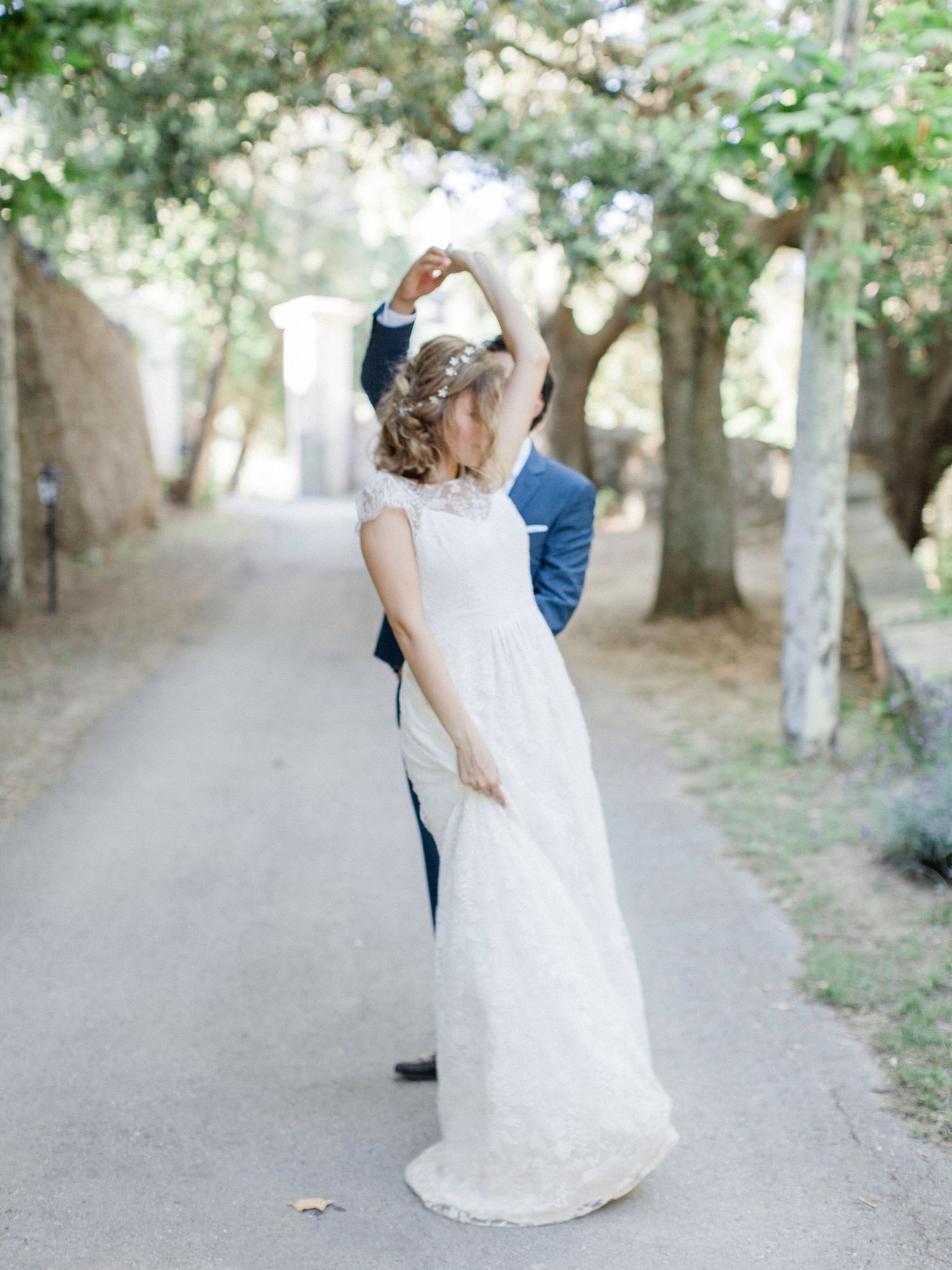 Bride in lace gown twirls with groom in blue suit on tree-lined path during outdoor wedding dance.
