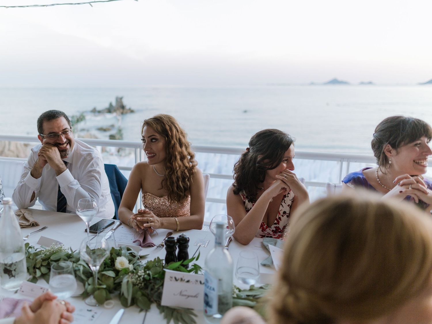 Des invités de mariage attablés lors d'une réception en plein air, avec une vue pittoresque sur l'océan au crépuscule.