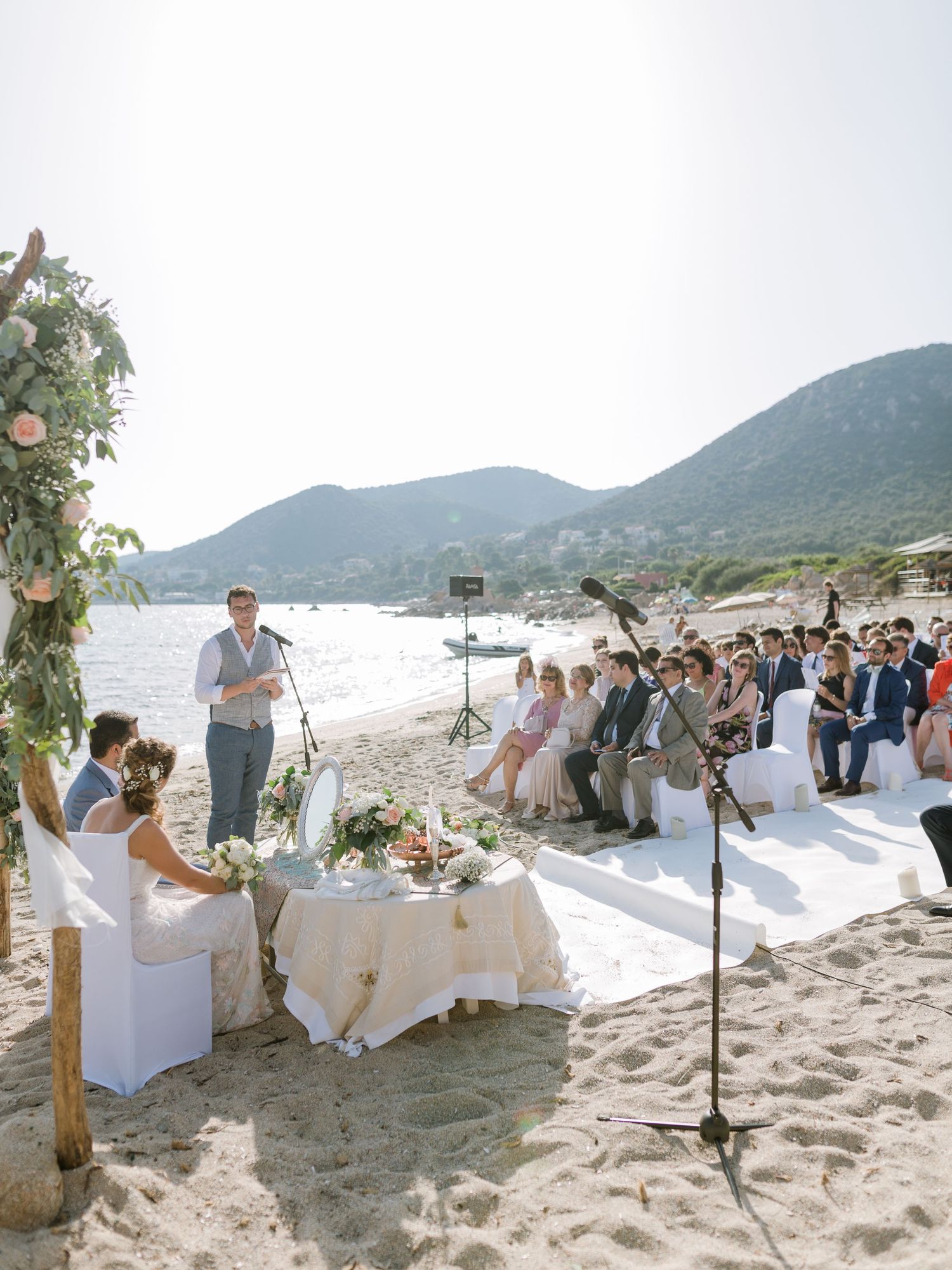 Cérémonie de mariage en plein air sur la plage, avec les invités assis au bord de la mer, une arche fleurie et les montagnes en arrière-plan