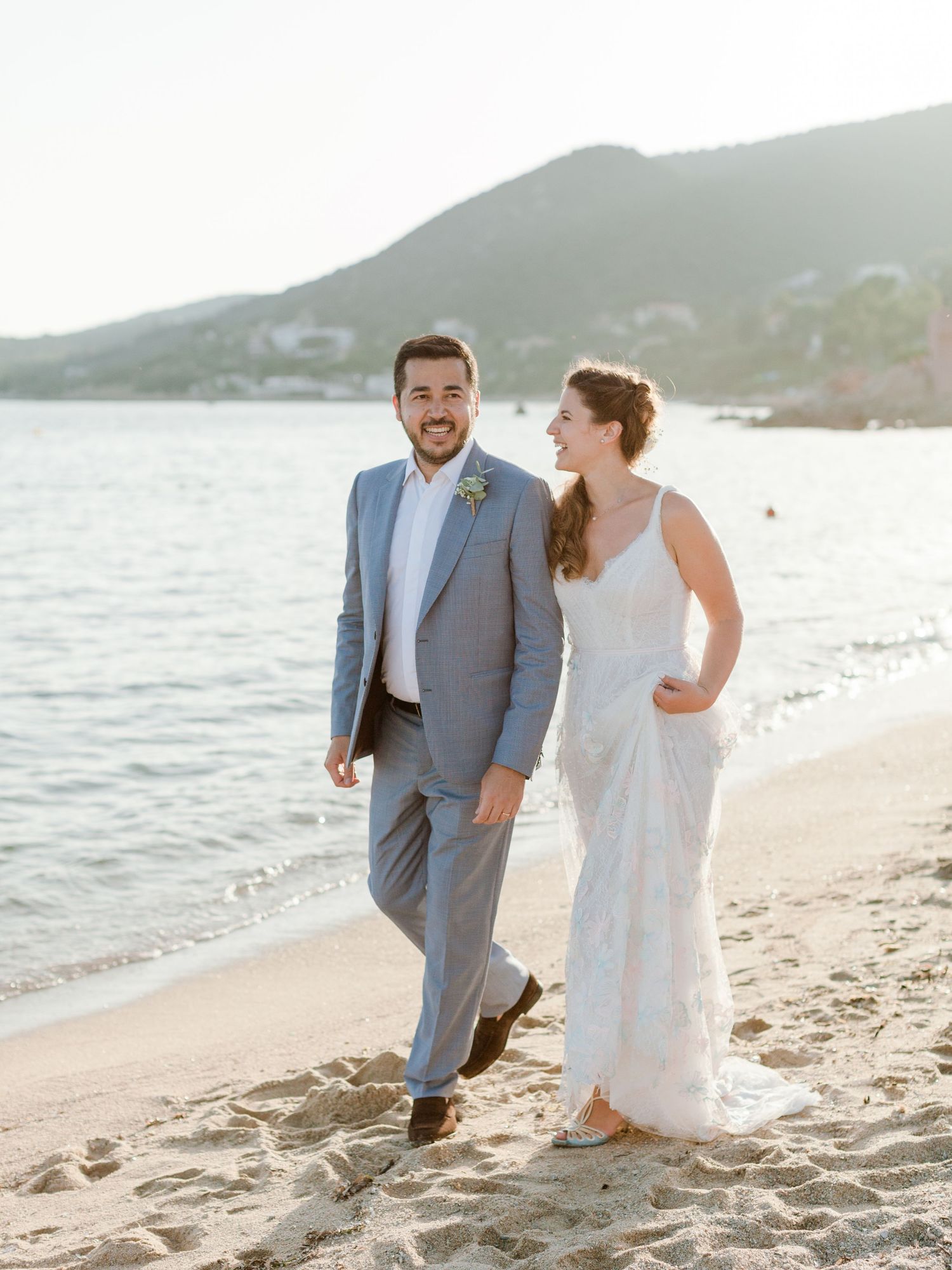 Les mariés marchent sur une plage de sable au coucher du soleil ; elle porte une robe blanche et lui un costume gris