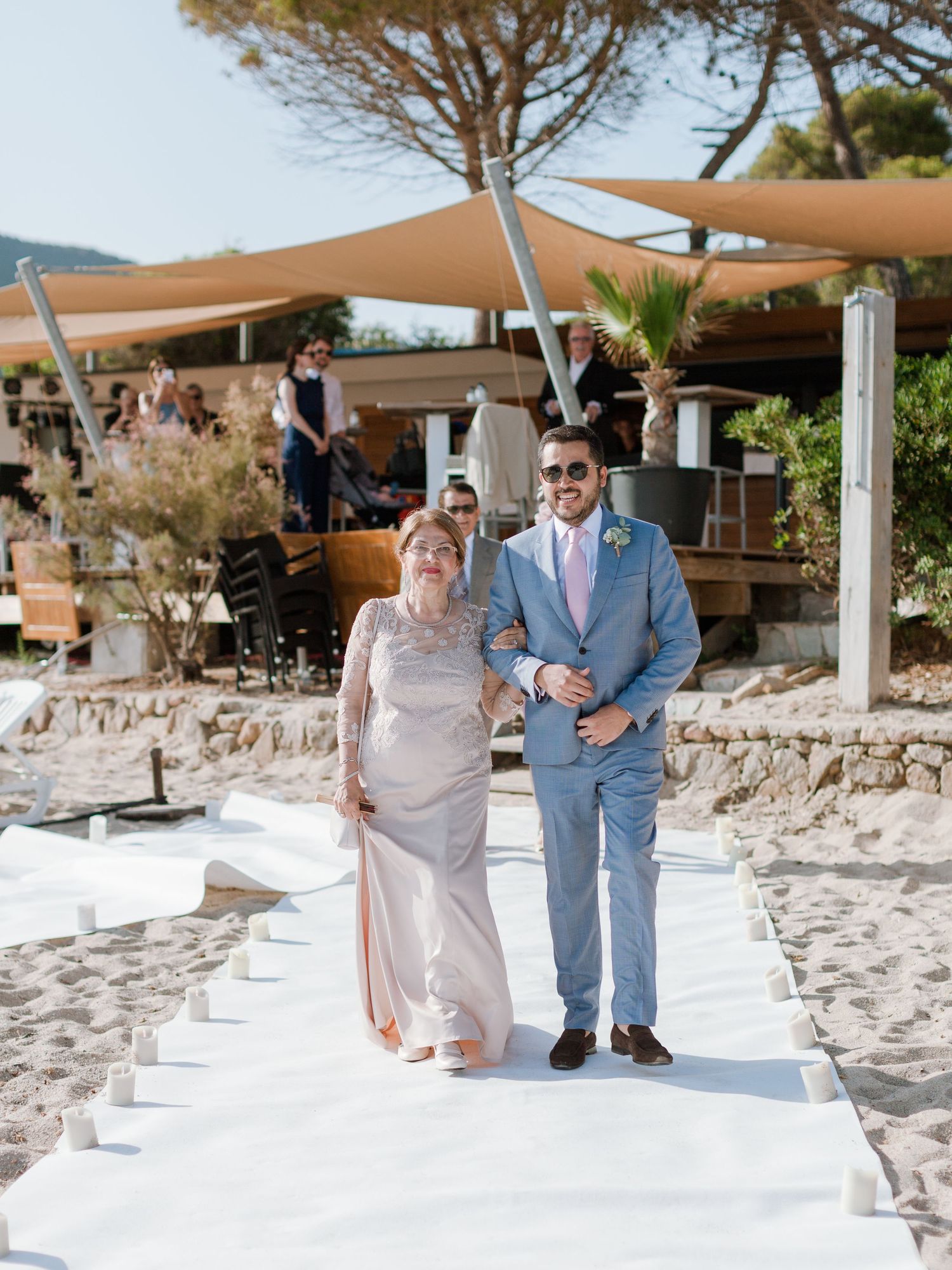 Un marié en costume bleu accompagne la mariée, vêtue d'une robe rose poudré, le long de l'allée blanche sur le sable d'un lieu de mariage