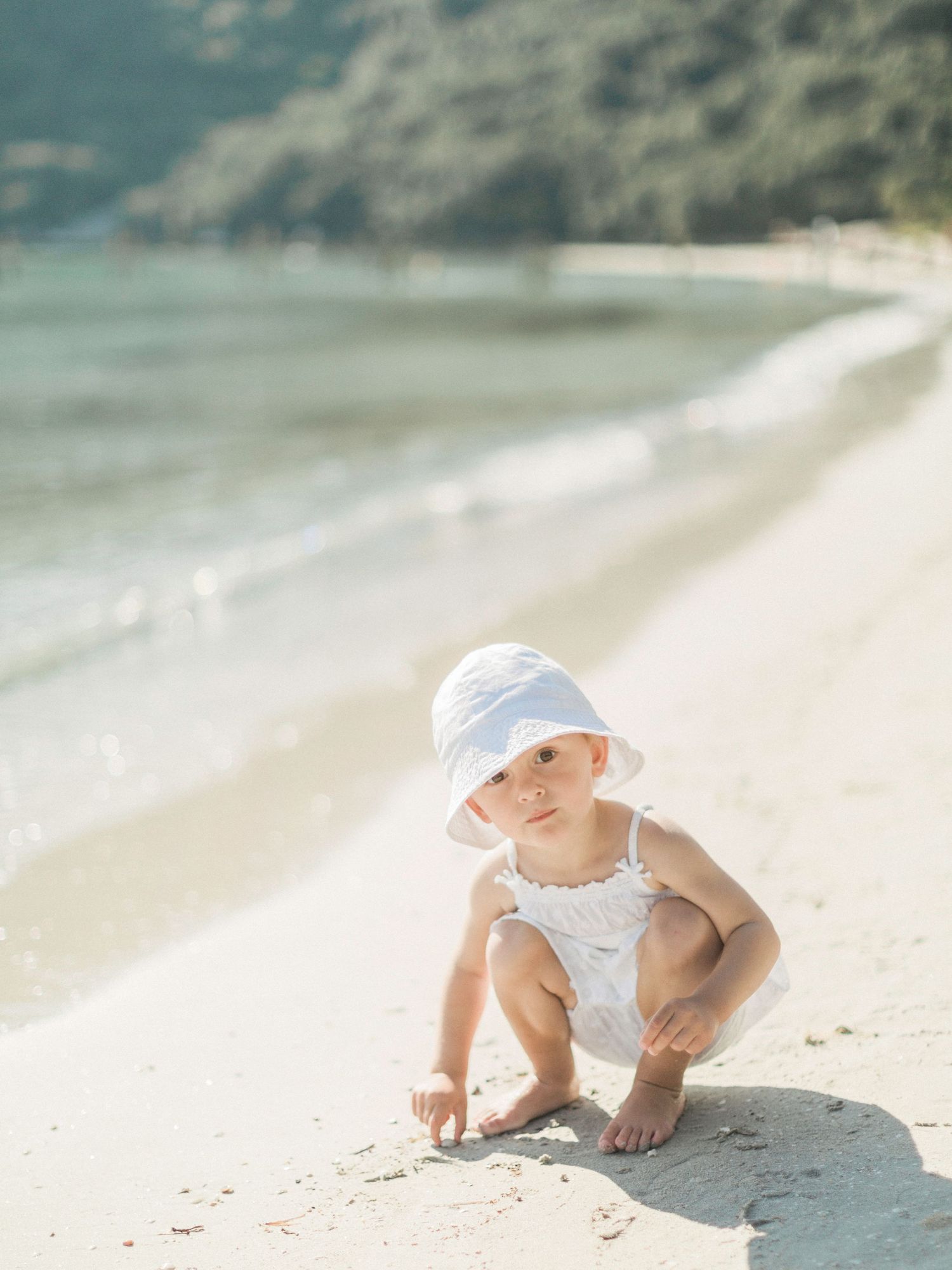 Un tout-petit coiffé d'un chapeau blanc est accroupi sur une plage de sable fin, près des douces vagues de l'océan. Plage de Santa Giulia.