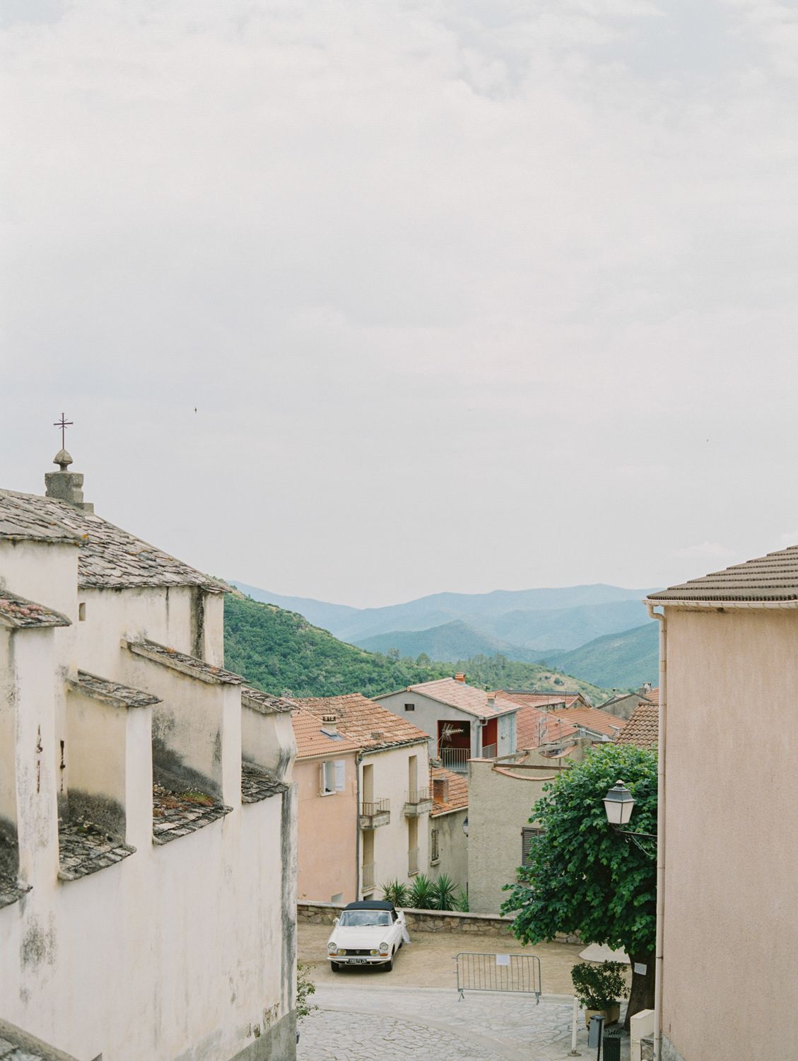 Charming Italian hillside village with terracotta rooftops, a vintage white car on cobblestone street, church cross, and green mountains.