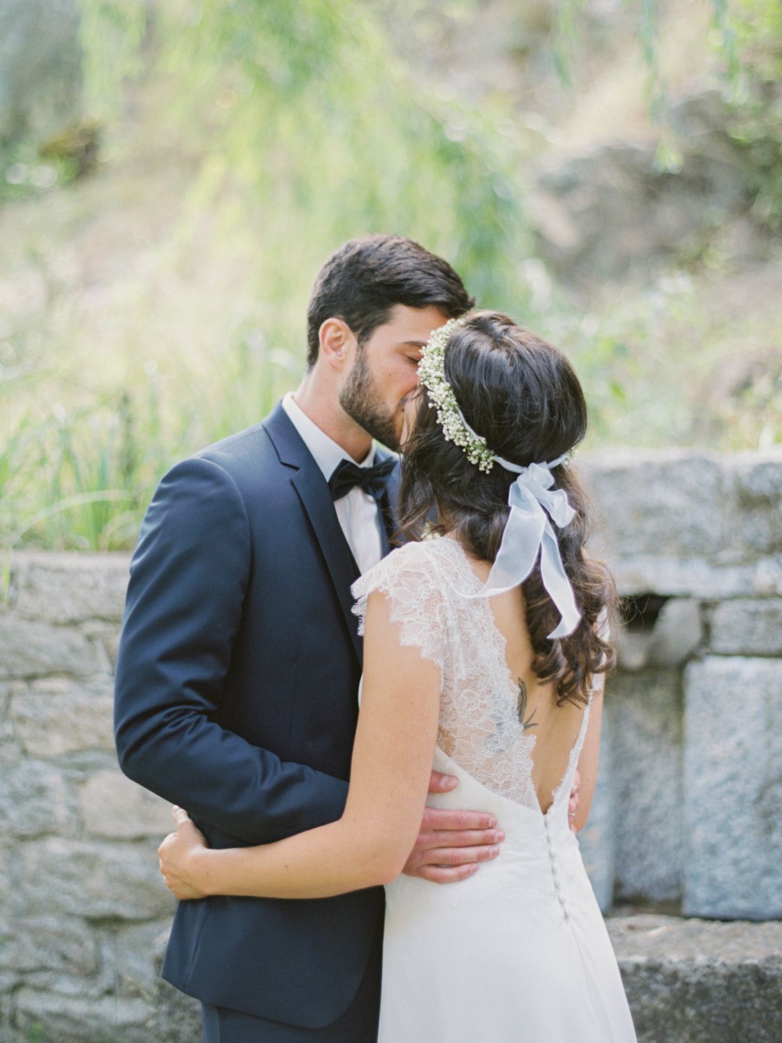 A bride and groom share a romantic kiss outdoors, she wears a lace gown with floral headpiece, he wears a navy tuxedo.
