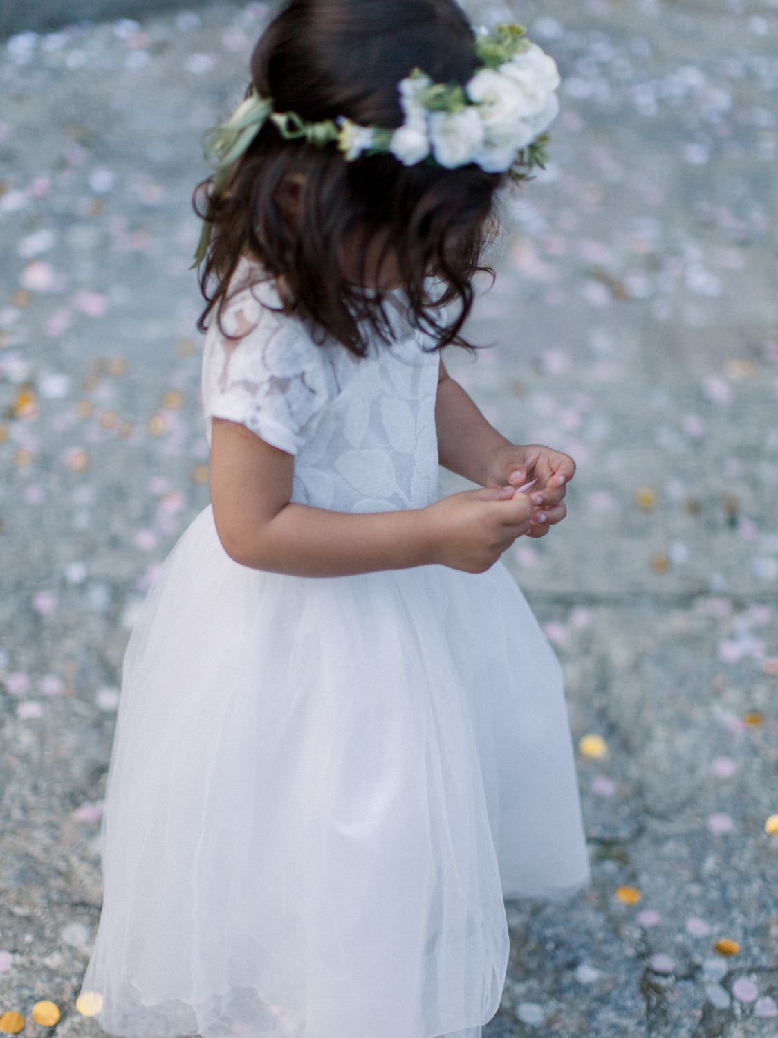Young flower girl in white tulle dress and floral crown stands on petal-strewn ground at a wedding.