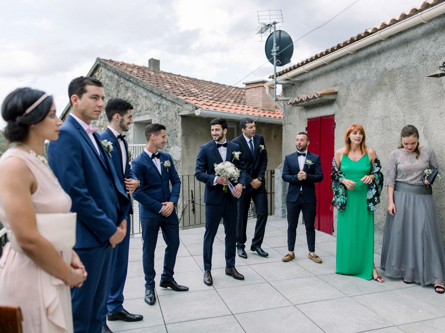 Wedding party gathered outdoors near rustic stone building, groomsmen in navy suits with bride and bridesmaids in elegant attire.