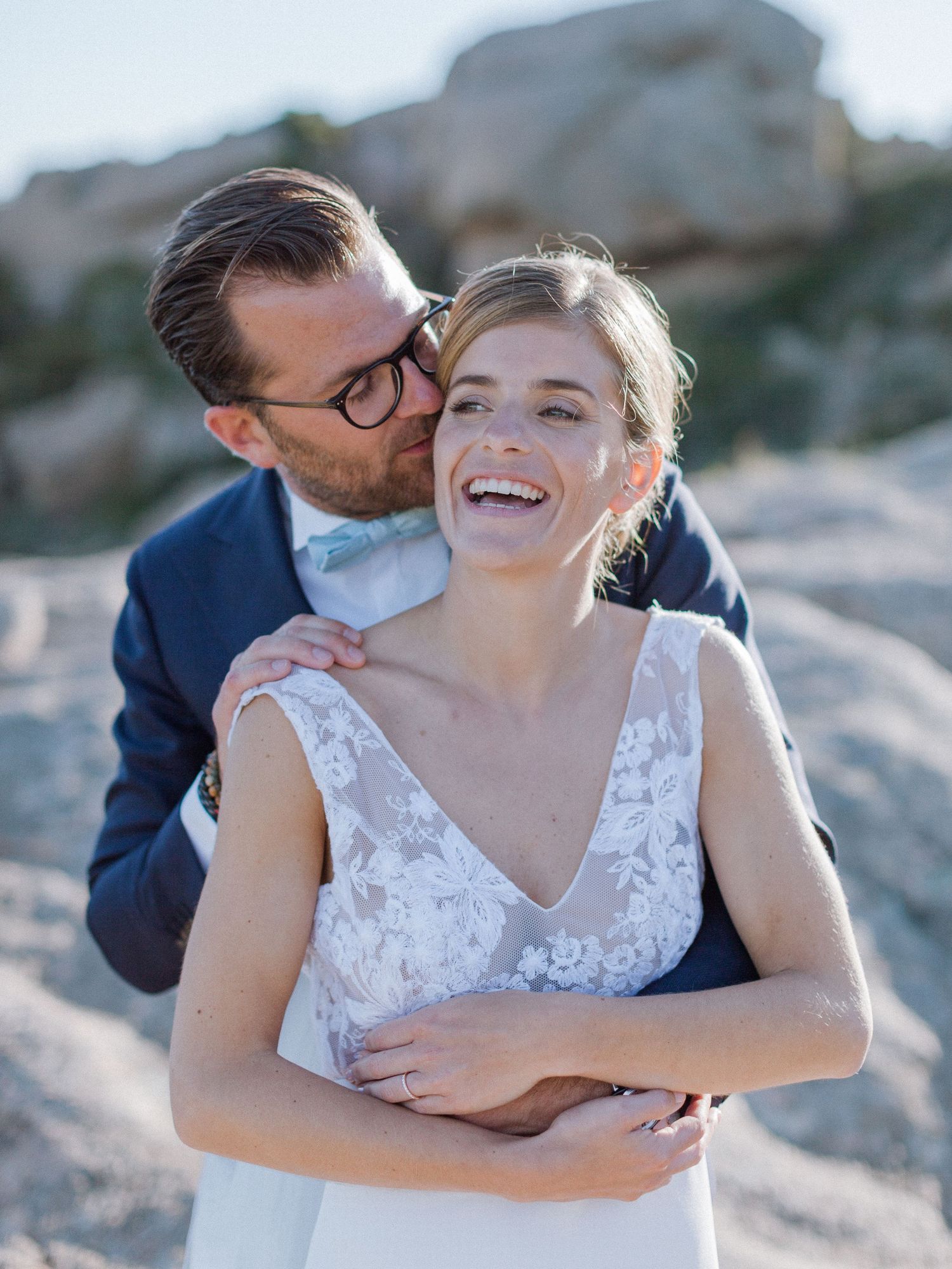 La mariée, radieuse en robe de dentelle, rit tandis que le marié en costume bleu marine l'enlace par derrière sur le lieu de leur mariage
