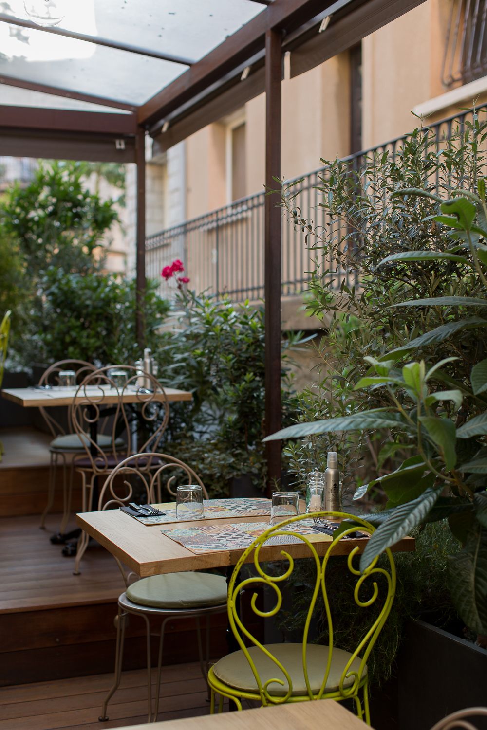 Charmante terrasse de restaurant en plein air avec des chaises en fer forgé jaunes et beiges, des tables en bois et une végétation luxuriant