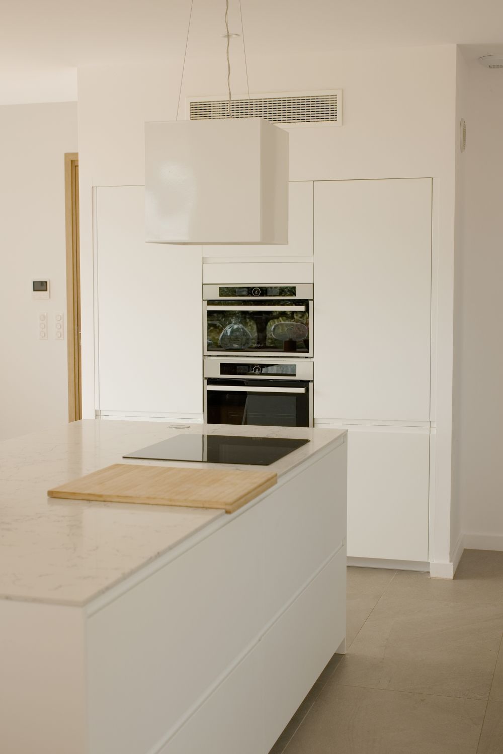Cuisine blanche moderne et minimaliste avec îlot central, fours encastrés, plaque à induction et plans de travail en marbre.