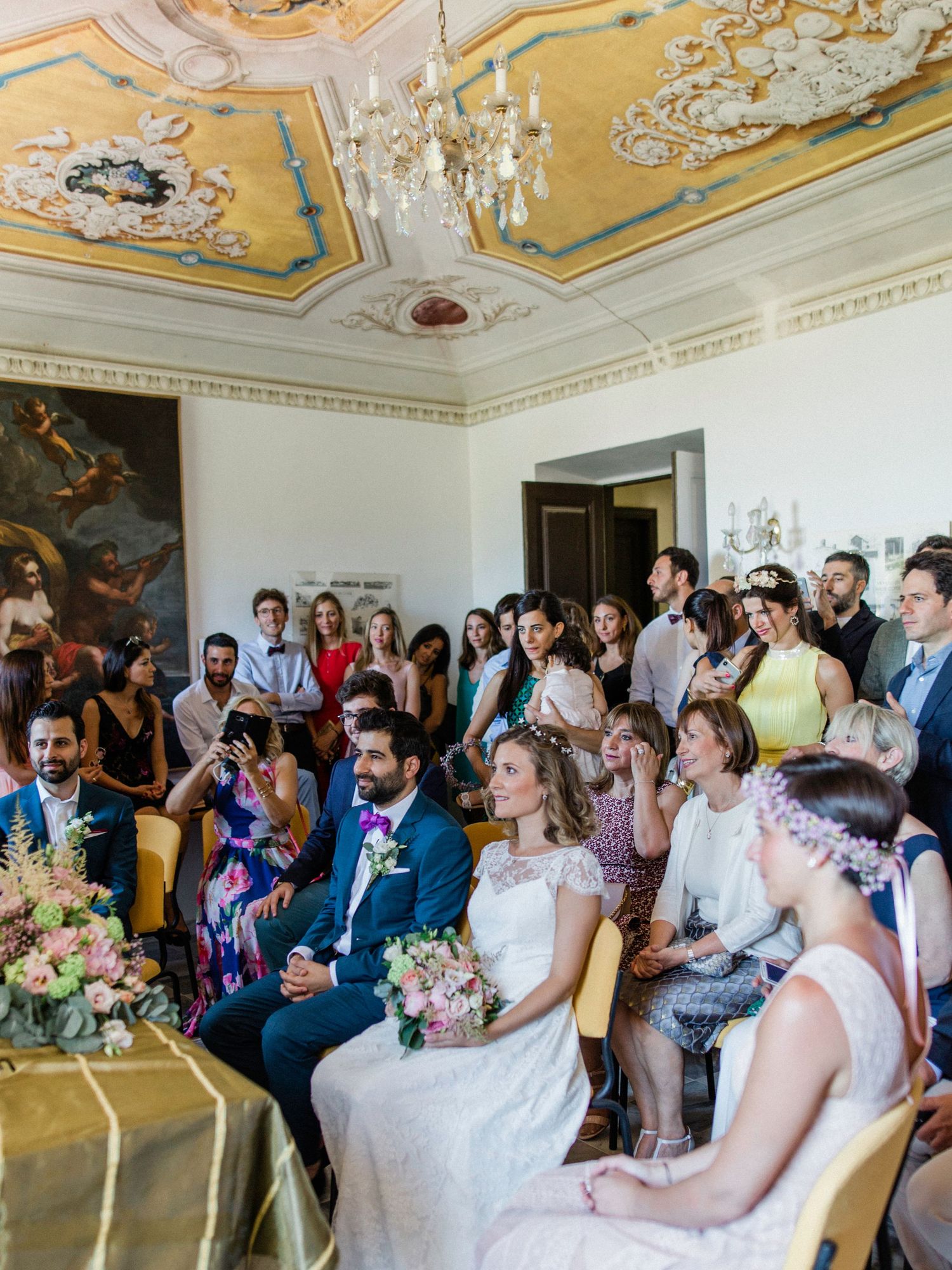 Elegant wedding ceremony in ornate room with gilded ceiling, bride and groom seated among guests holding bouquets.
