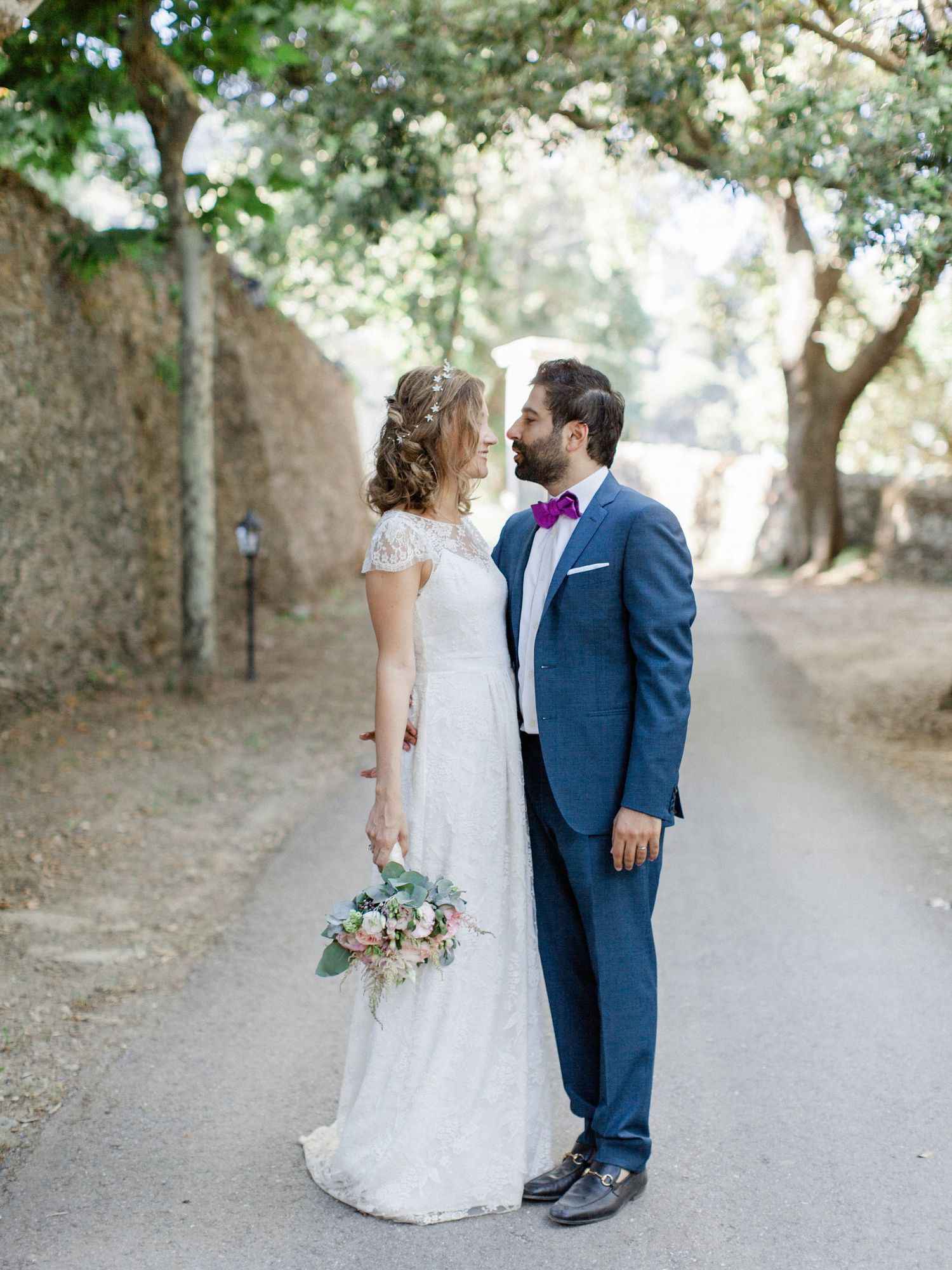 Bride and groom share a romantic moment on a tree-lined path, she holds a bouquet, he wears a blue suit with purple bow tie.