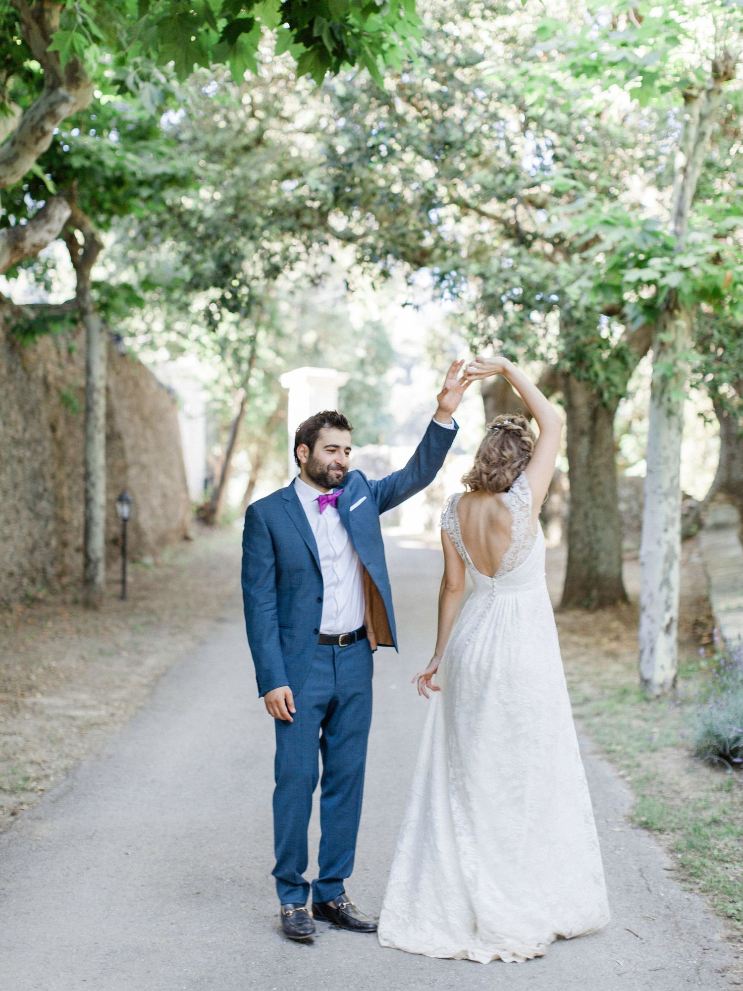 Groom in blue suit twirling bride in white backless gown on tree-lined path at outdoor wedding.
