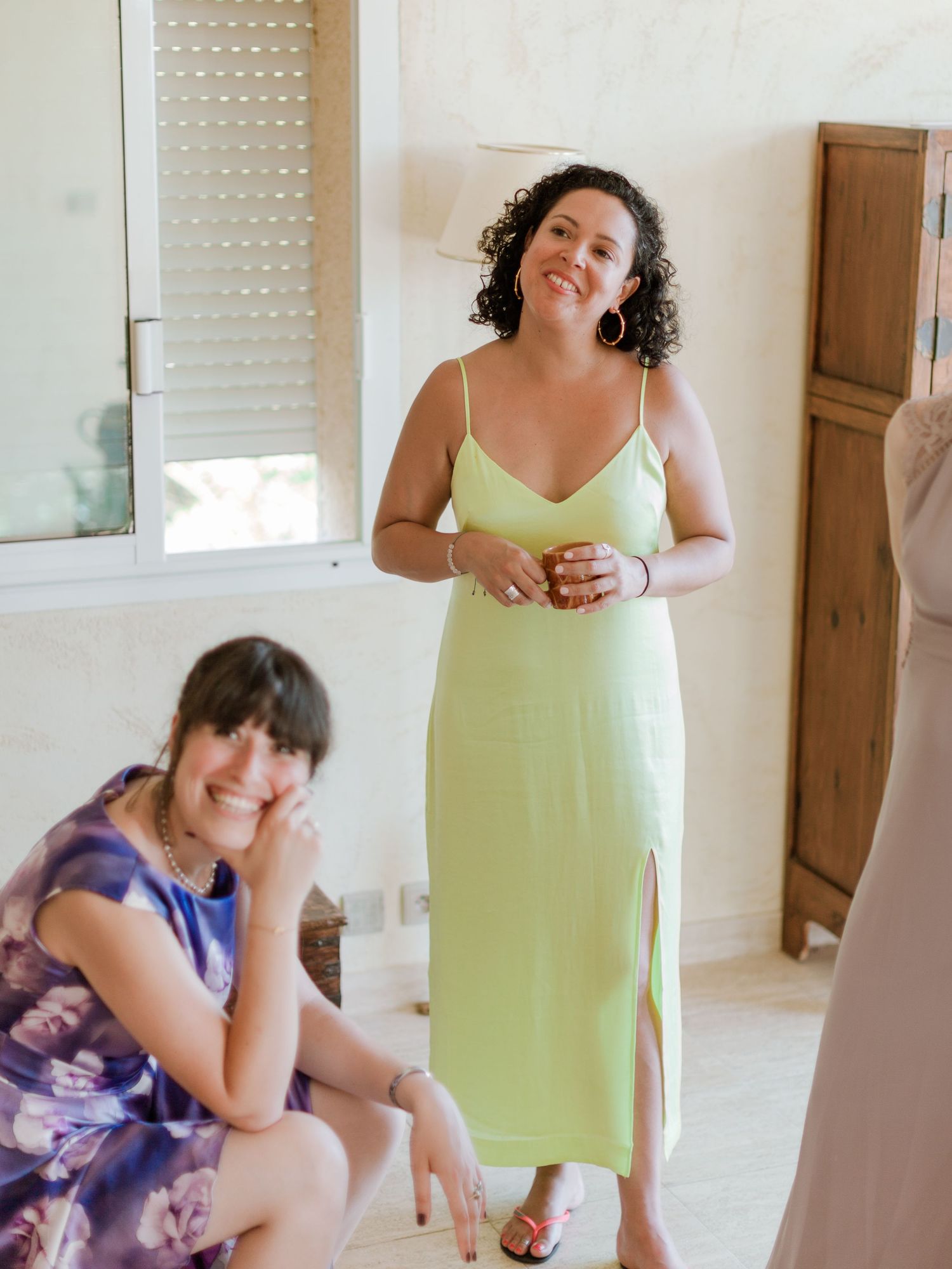 Deux femmes dans une pièce lumineuse : l'une porte une robe nuisette verte et tient une boisson, l'autre est assise et porte une robe violet