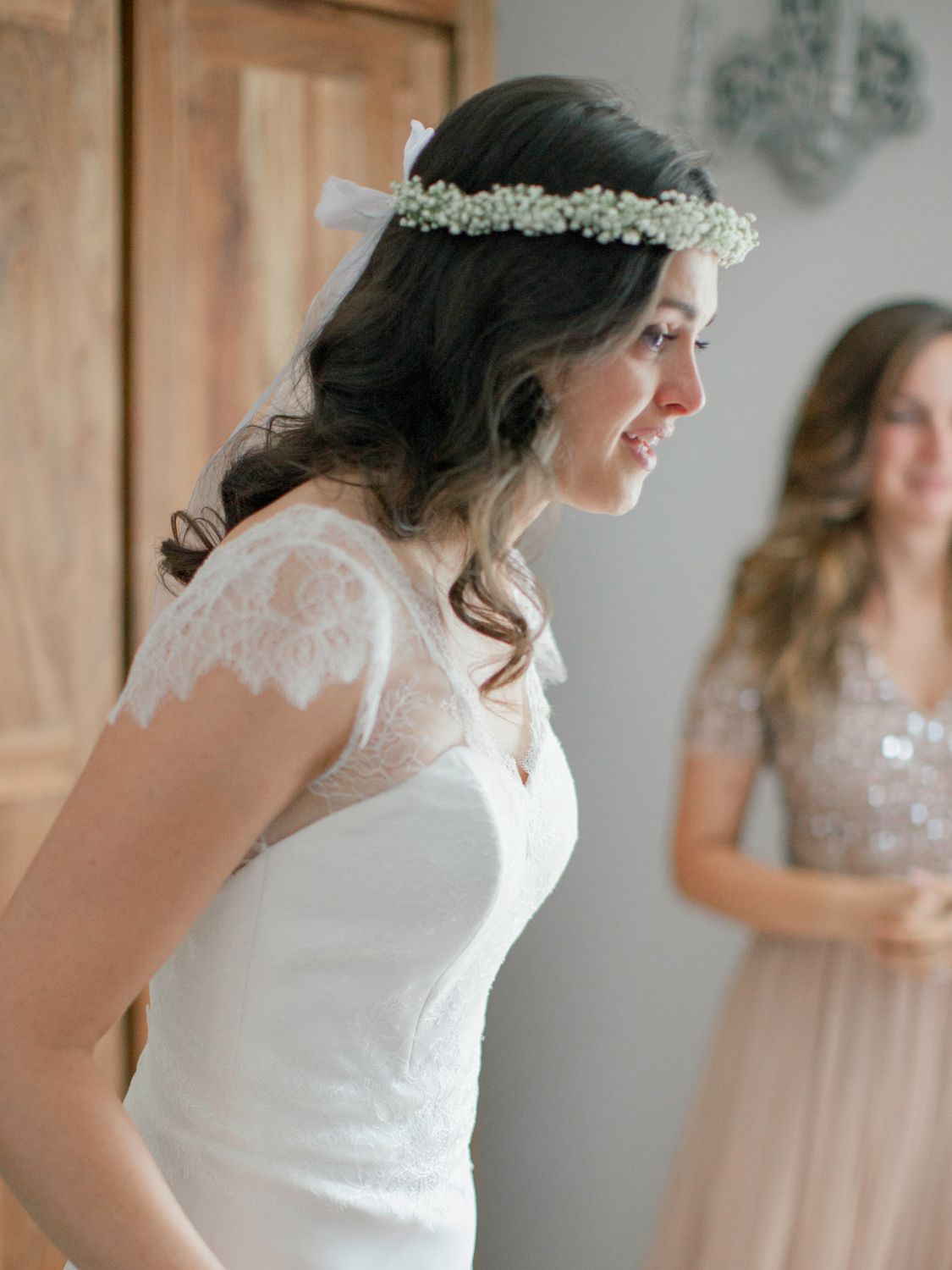 Bride in white lace wedding dress and floral crown smiles softly, with bridesmaid in blush gown visible behind her.