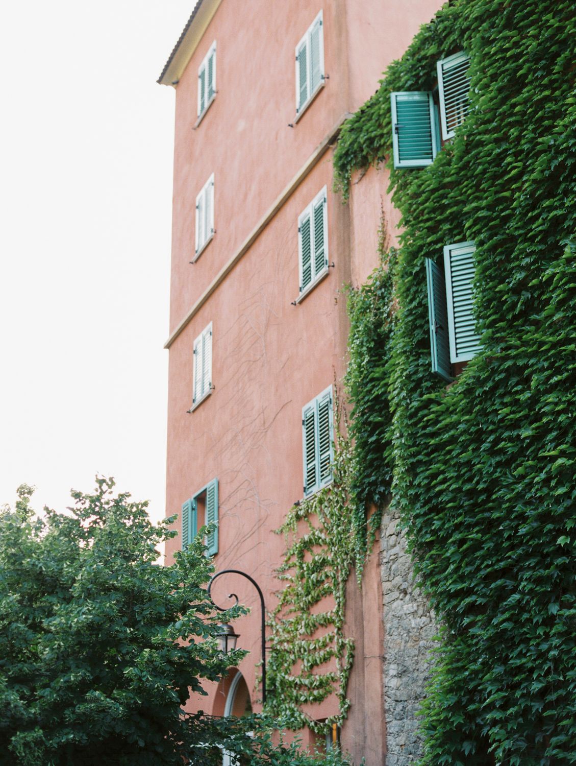 Pink building covered in lush green ivy with teal shutters and windows in a charming European village setting.