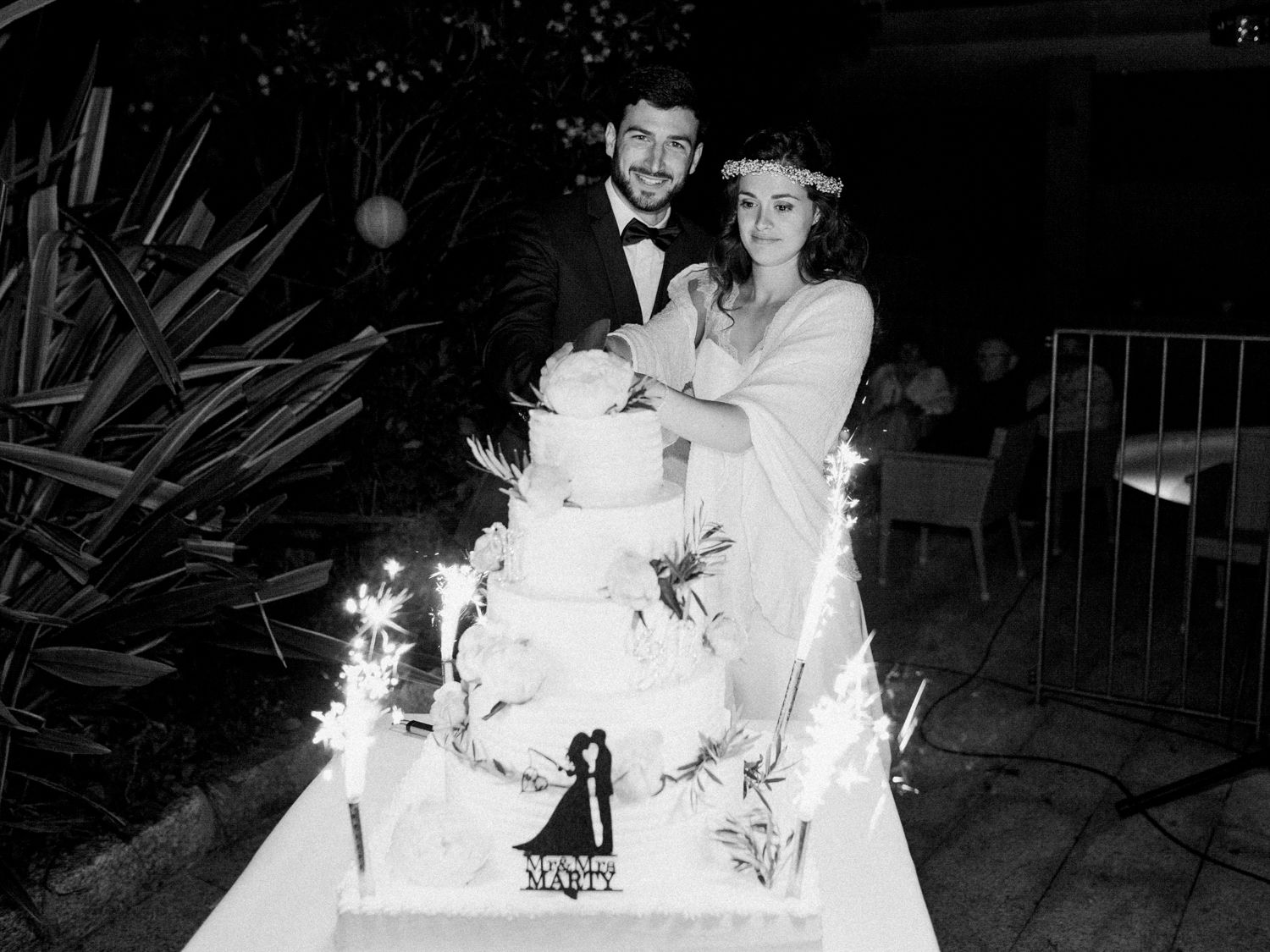Photo en noir et blanc de jeunes mariés coupant leur gâteau de mariage avec des cierges magiques lors d'une réception nocturne en plein air.