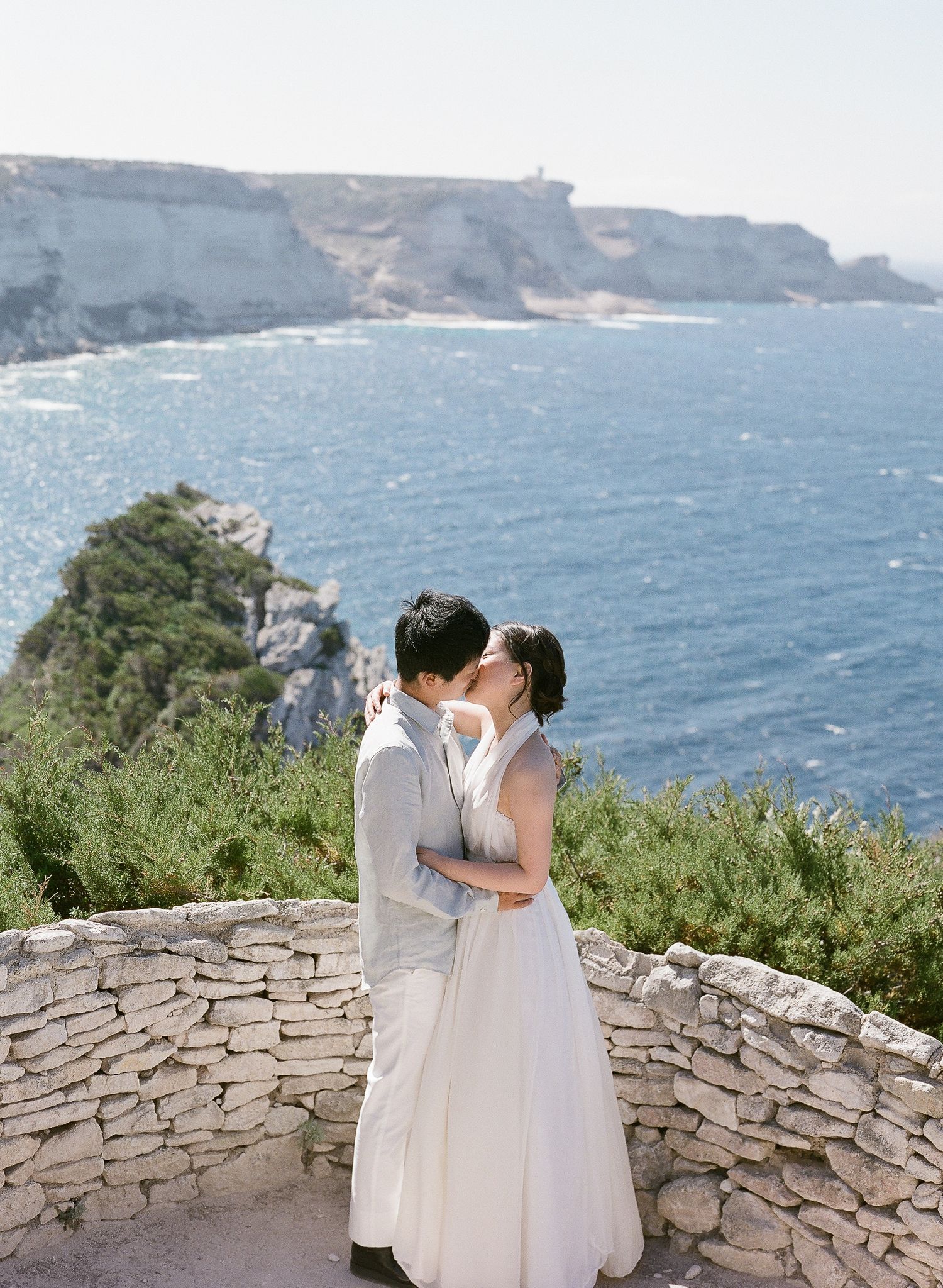 Des mariés asiatiques échangent un baiser sur un muret de pierre, avec des falaises côtières spectaculaires et la mer bleue en arrière-plan.