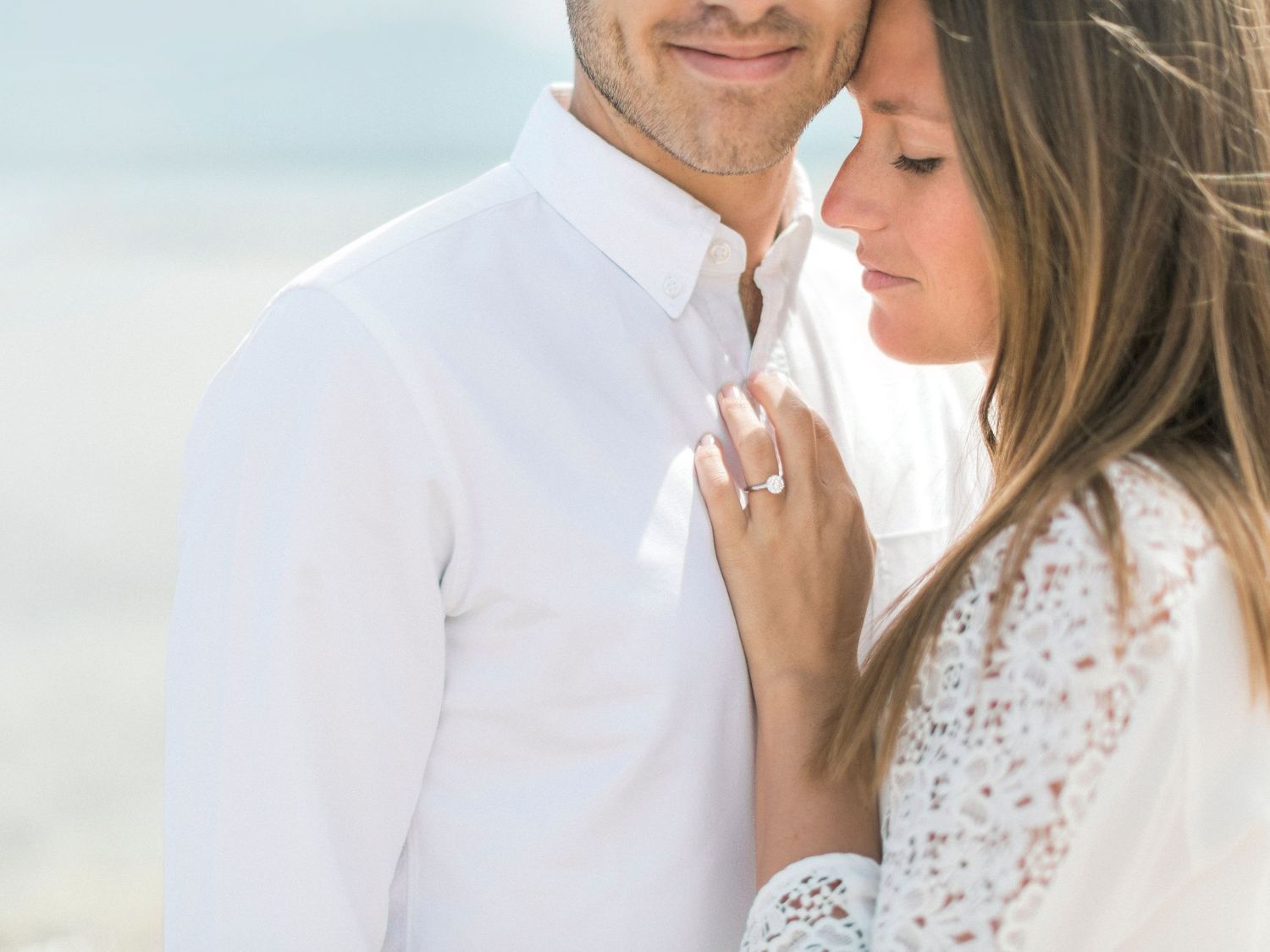 Gros plan sur un couple souriant en tenues blanches, s'enlaçant sur une plage ensoleillée ; la femme porte une bague. Plage de Calvi.