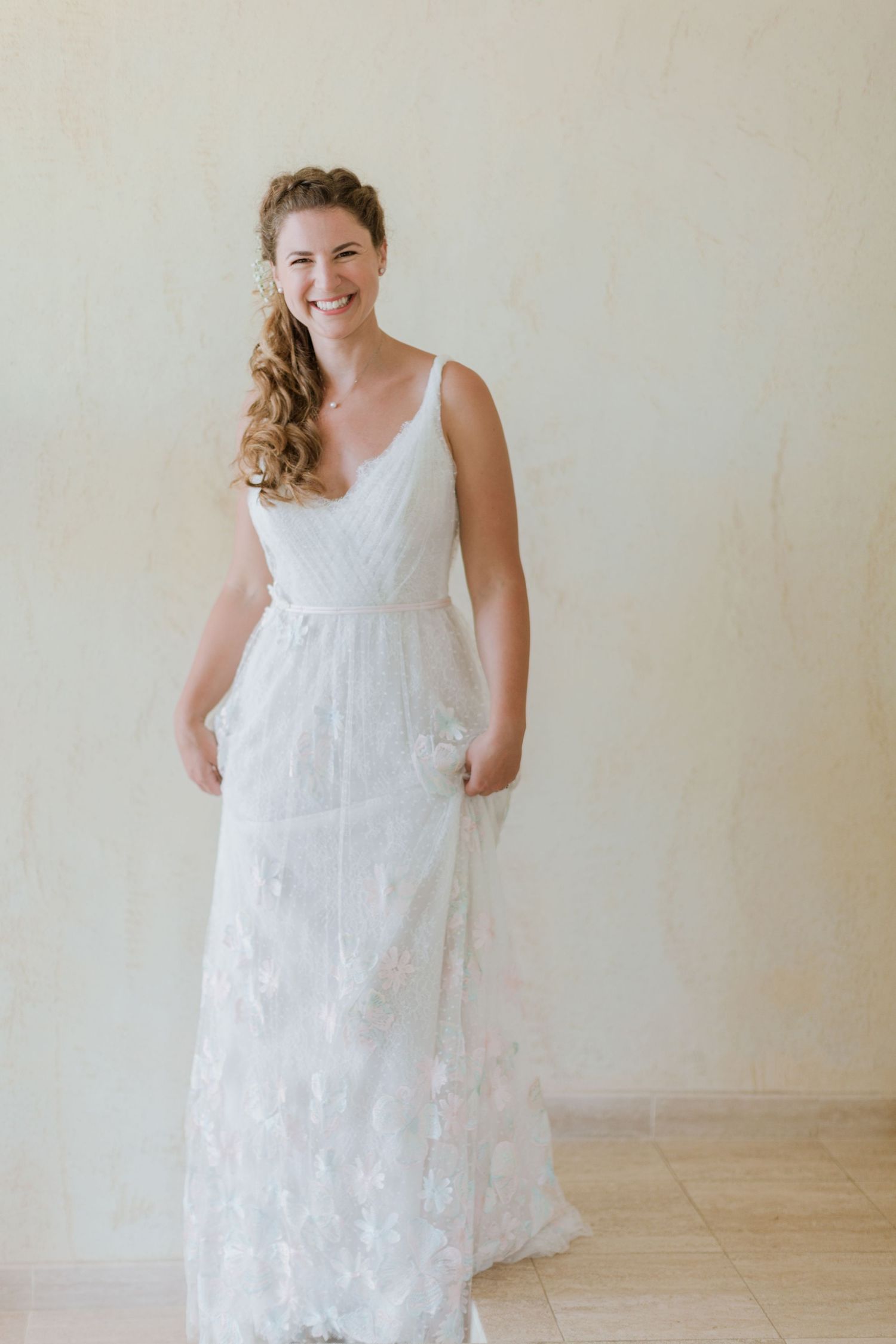 Une mariée radieuse, vêtue d'une robe de mariée en dentelle florale et coiffée d'une tresse relevée, sourit sur un fond neutre.