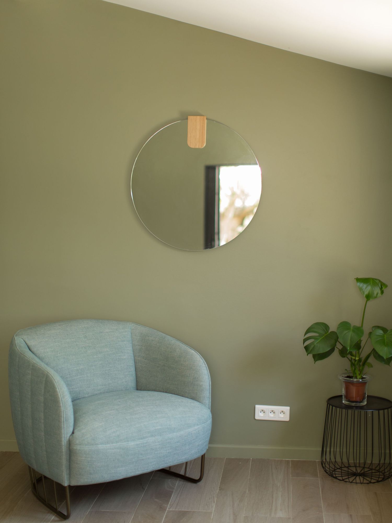 Espace de vie cosy avec un fauteuil bleu clair, un miroir rond et une plante monstera sur un mur vert. Prise de vue à Porto-Vecchio.