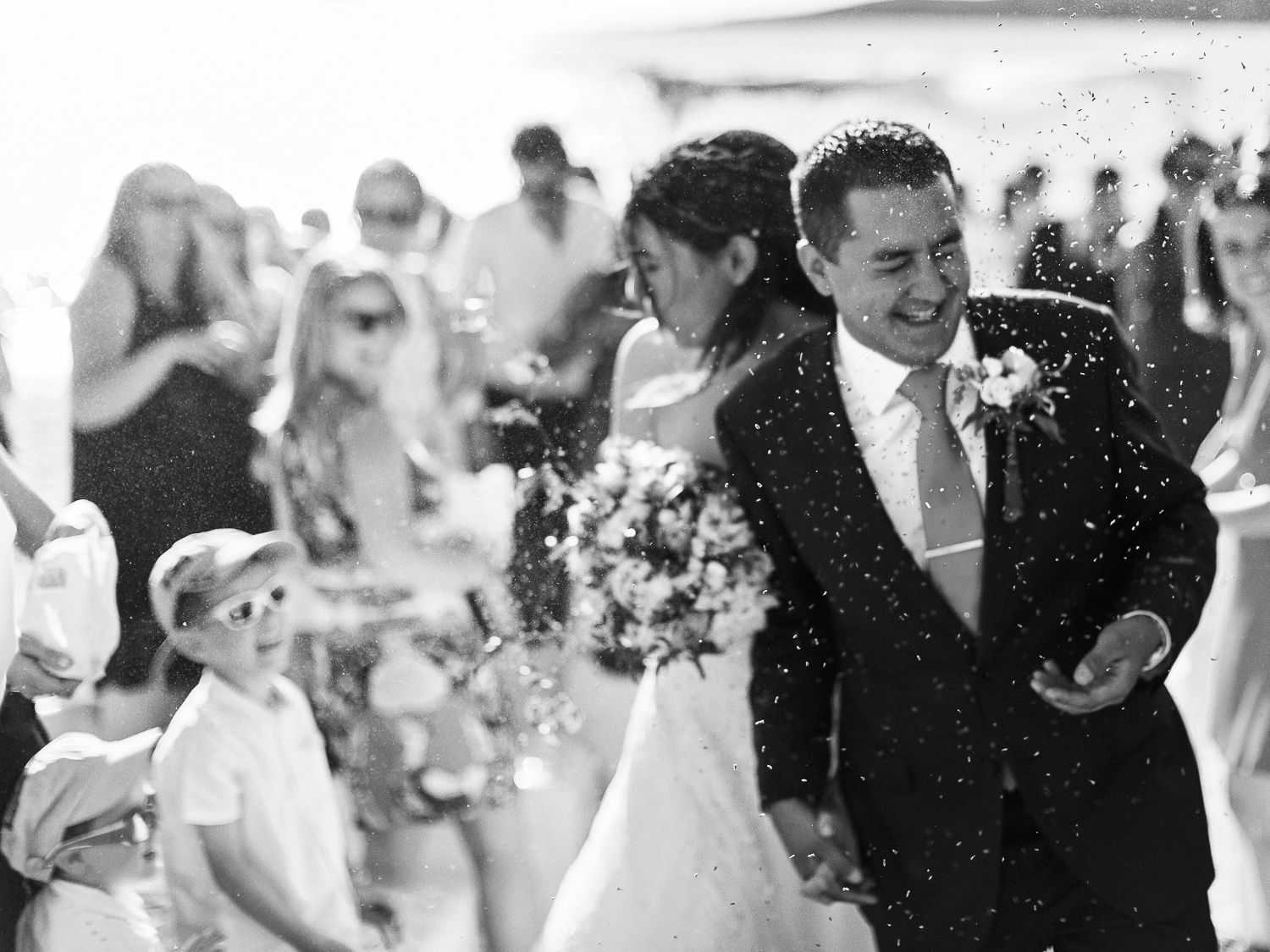 Photo en noir et blanc d'un couple de jeunes mariés rayonnants traversant une pluie de confettis, entourés de leurs invités.