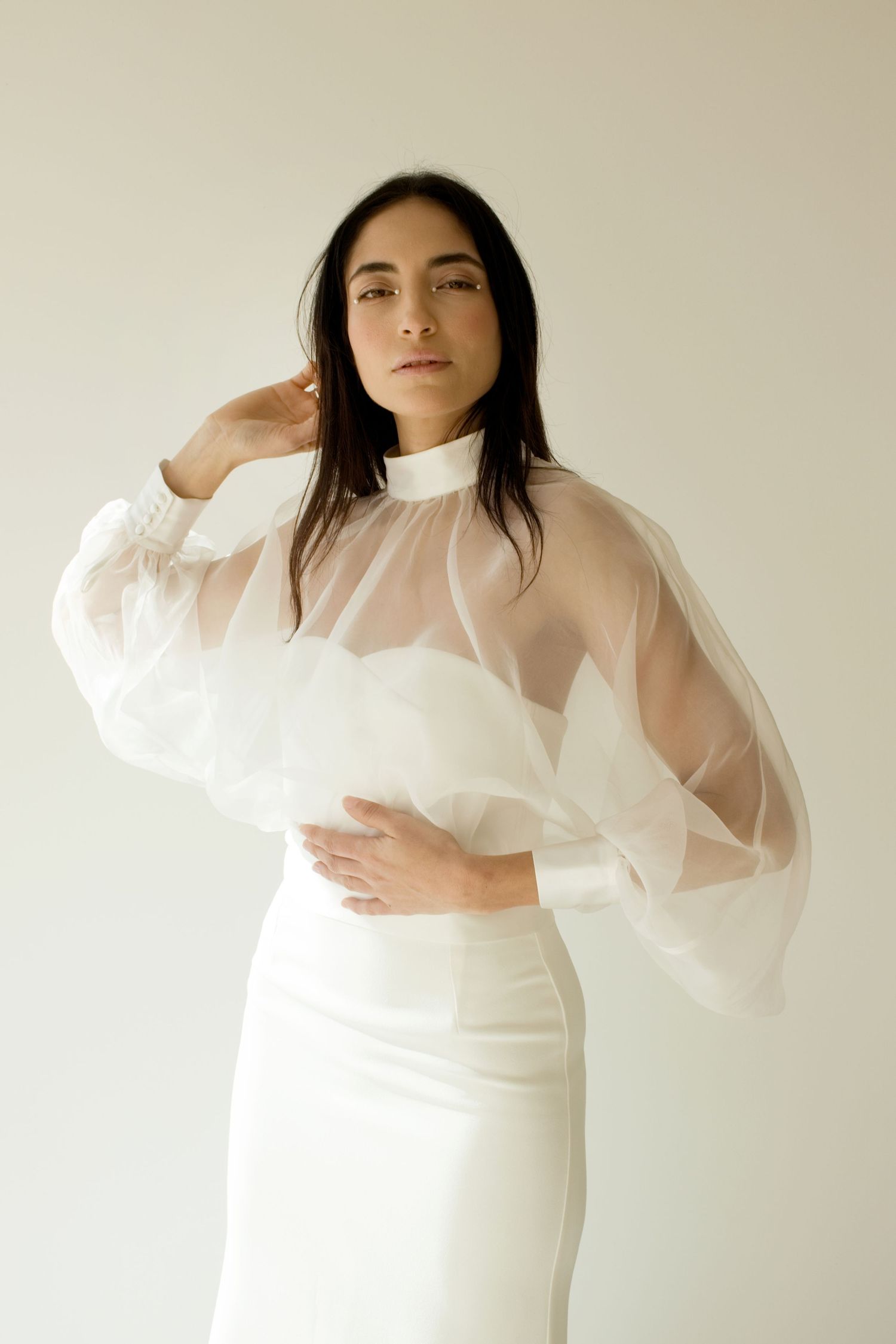 Une femme vêtue d'un haut en organza blanc by romance transparent à manches ballons et d'une jupe en satin blanc pose sur un fond clair.