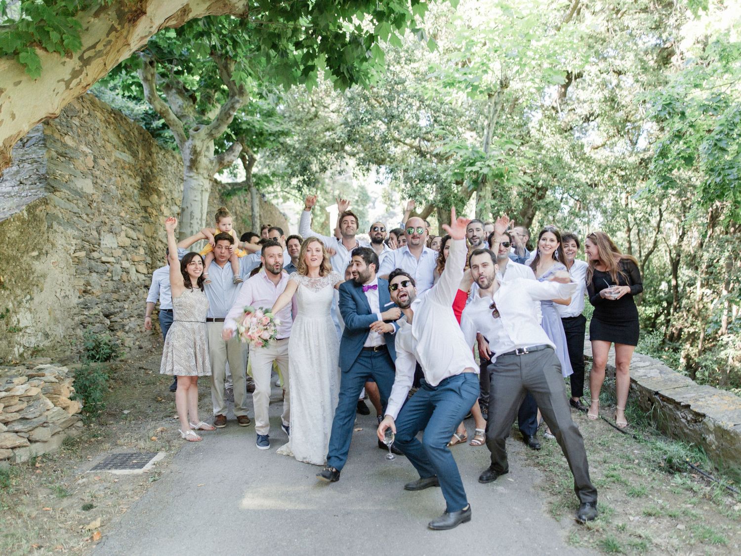 Wedding party celebrating outdoors under trees, guests cheering and posing playfully with bride and groom on stone path.